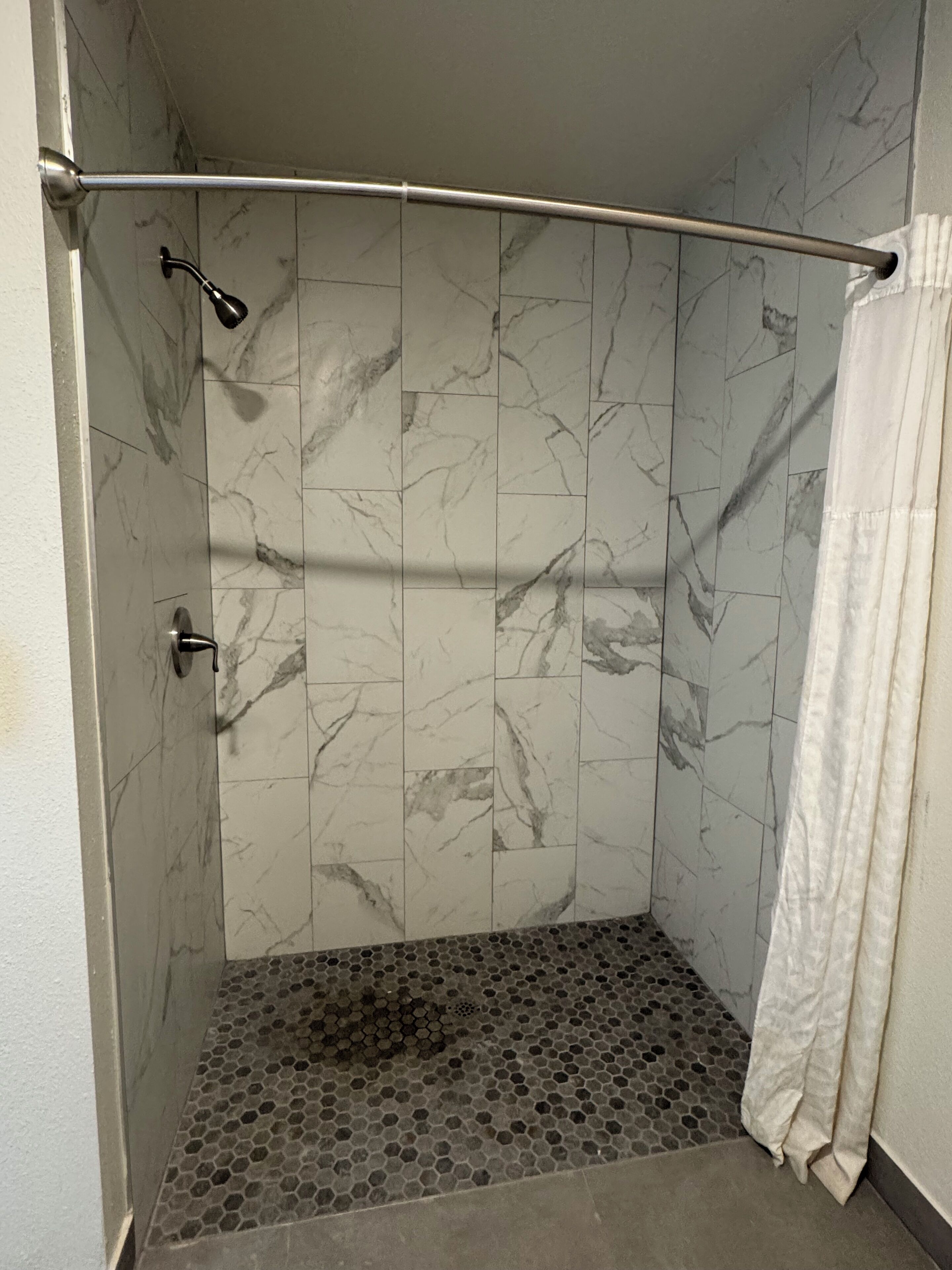 Ducha con barra metálica curva, suelo de azulejos hexagonales y paredes con diseño de mármol.