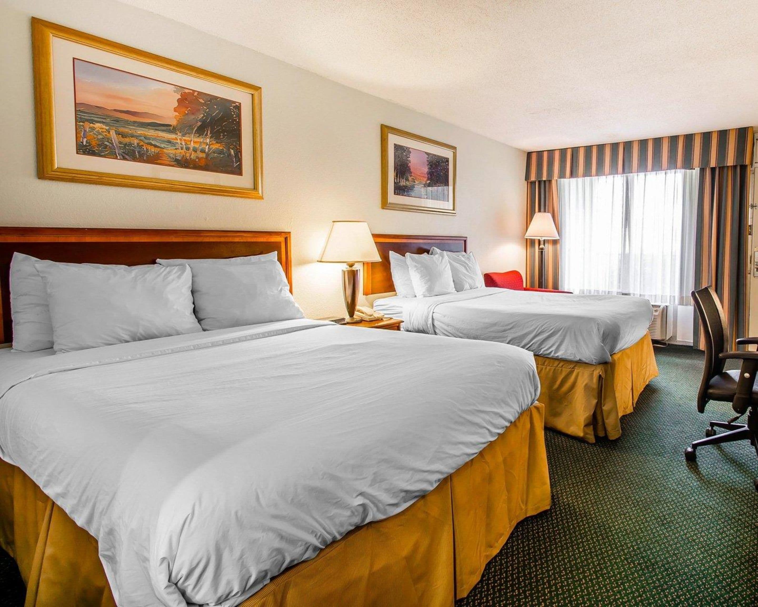 A hotel room with two beds, a wooden headboard, a desk with a lamp, a chair, and two framed pictures on the wall.