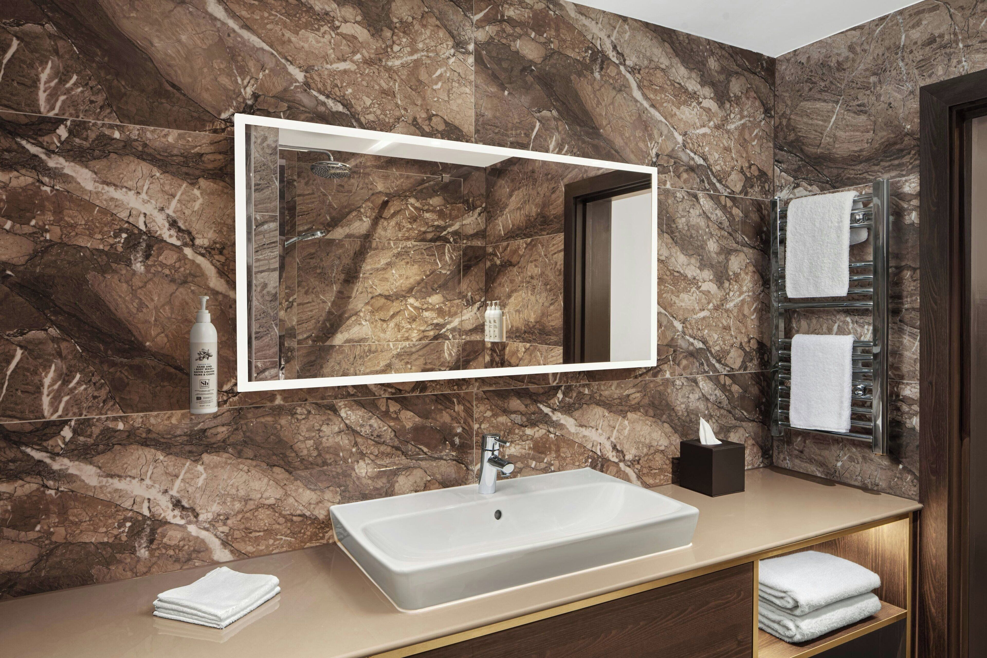A bathroom with a marble wall, a large mirror, a white sink, and a towel rack.