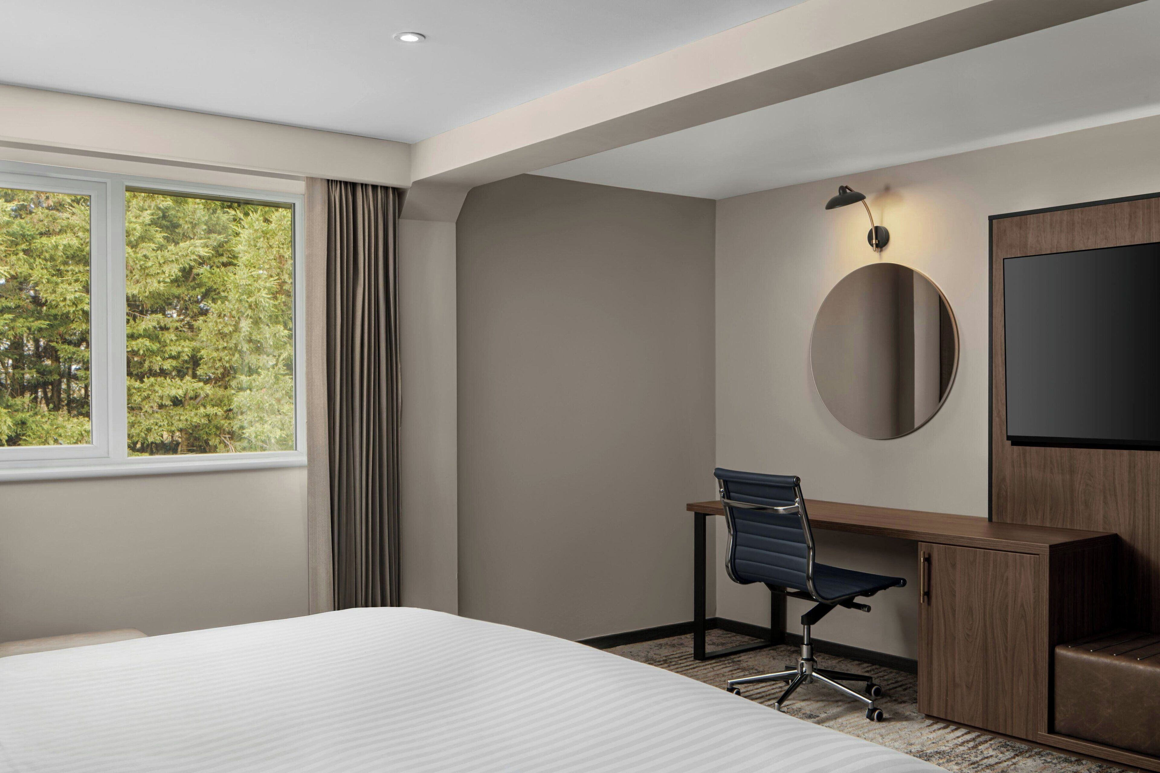 A hotel room with a bed, a desk with a chair, a TV, and a window with curtains.