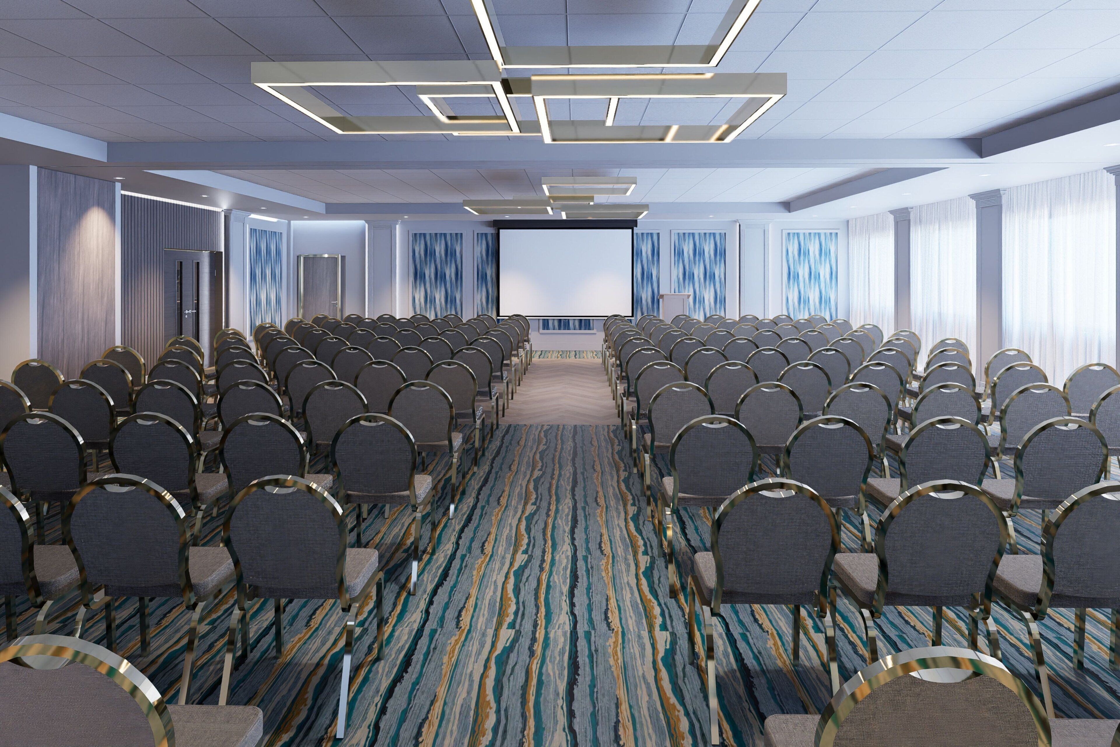 Un salón de conferencias con filas de sillas, una pantalla grande al frente y alfombra estampada.