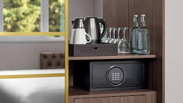 Een hotelkamer met een bed, een houten kastje met een kluisje, en diverse spullen zoals een waterkoker, kopjes en flessen.