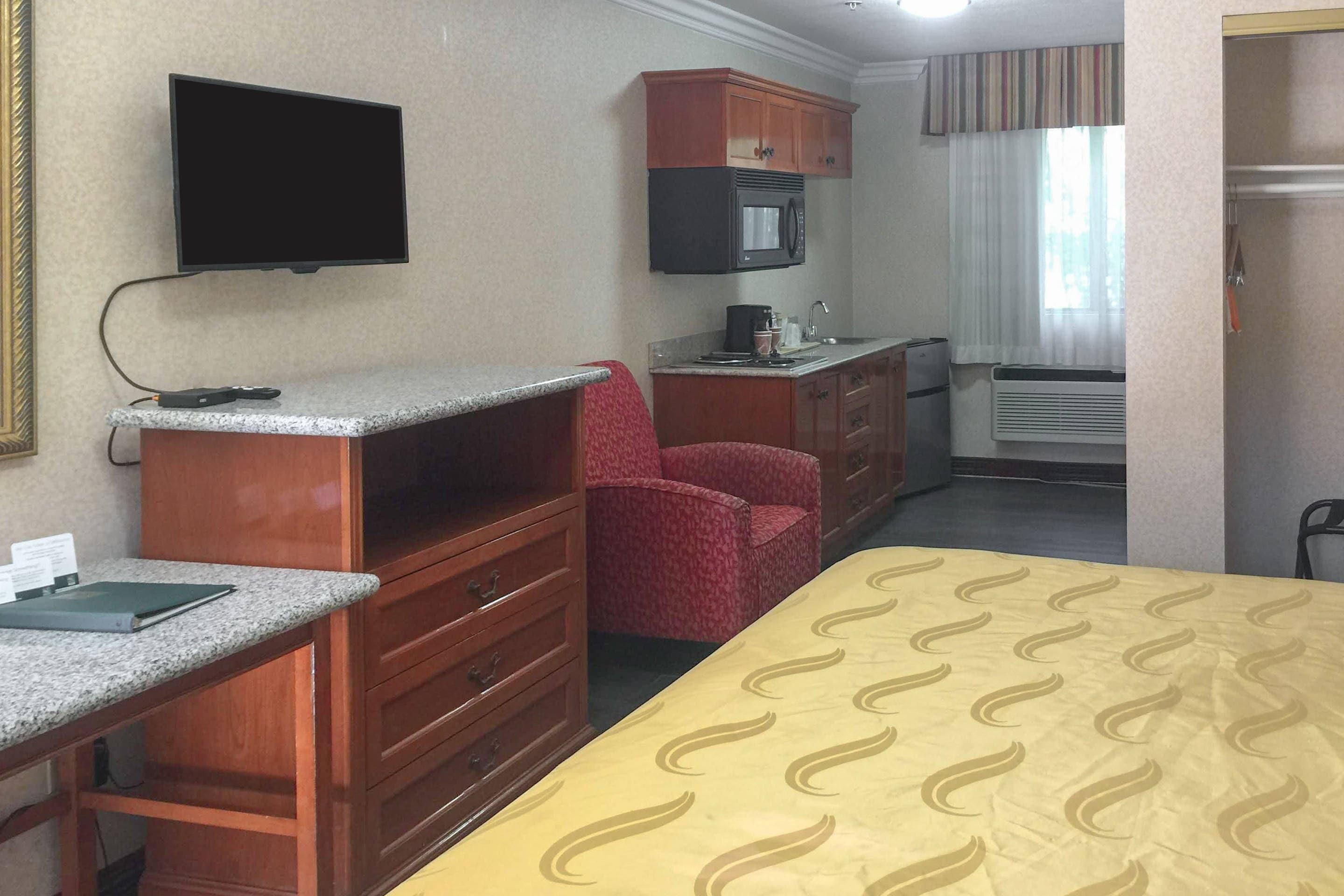 A hotel room with a bed, a desk with a flat-screen TV, a kitchenette with a microwave, a red chair, and a window with curtains.