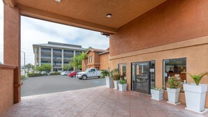 Entrada de hotel com alpendre coberto, plantas em vasos e estacionamento com vários carros.