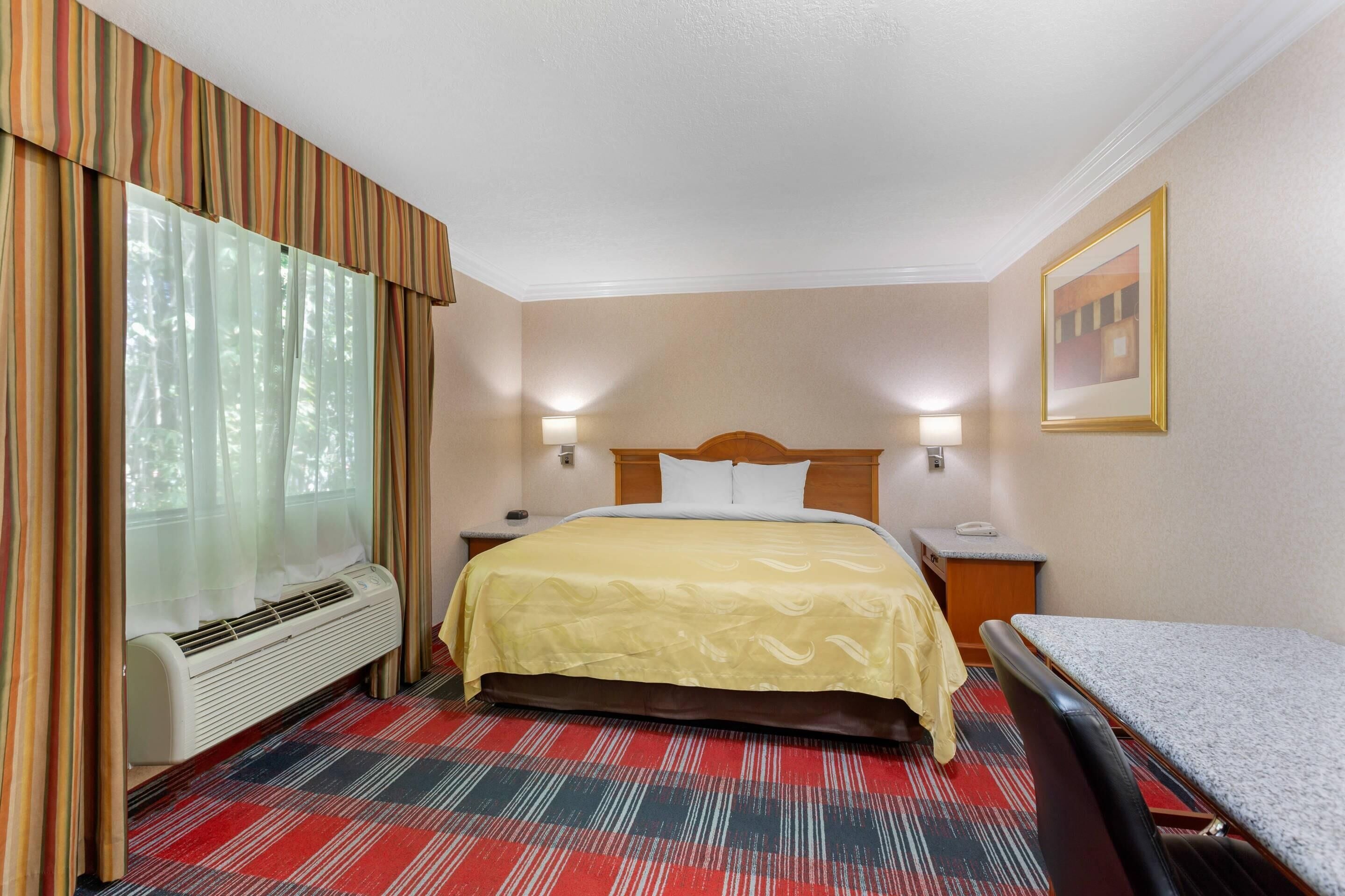 Habitación de hotel con cama, escritorio, silla, calefactor, ventana con cortinas e imagen colgada en la pared.