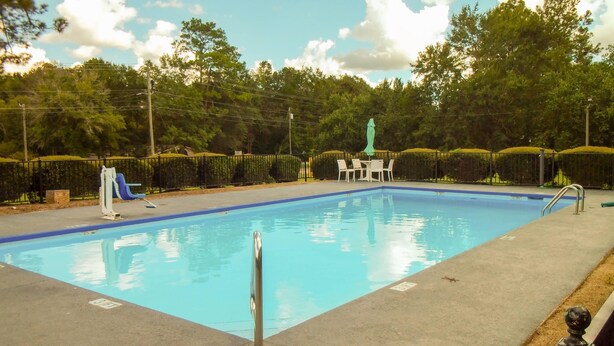 Piscina retangular com borda azul, rodeada por um deck pavimentado. Há cadeiras de descanso e uma mesa com guarda-sol verde.