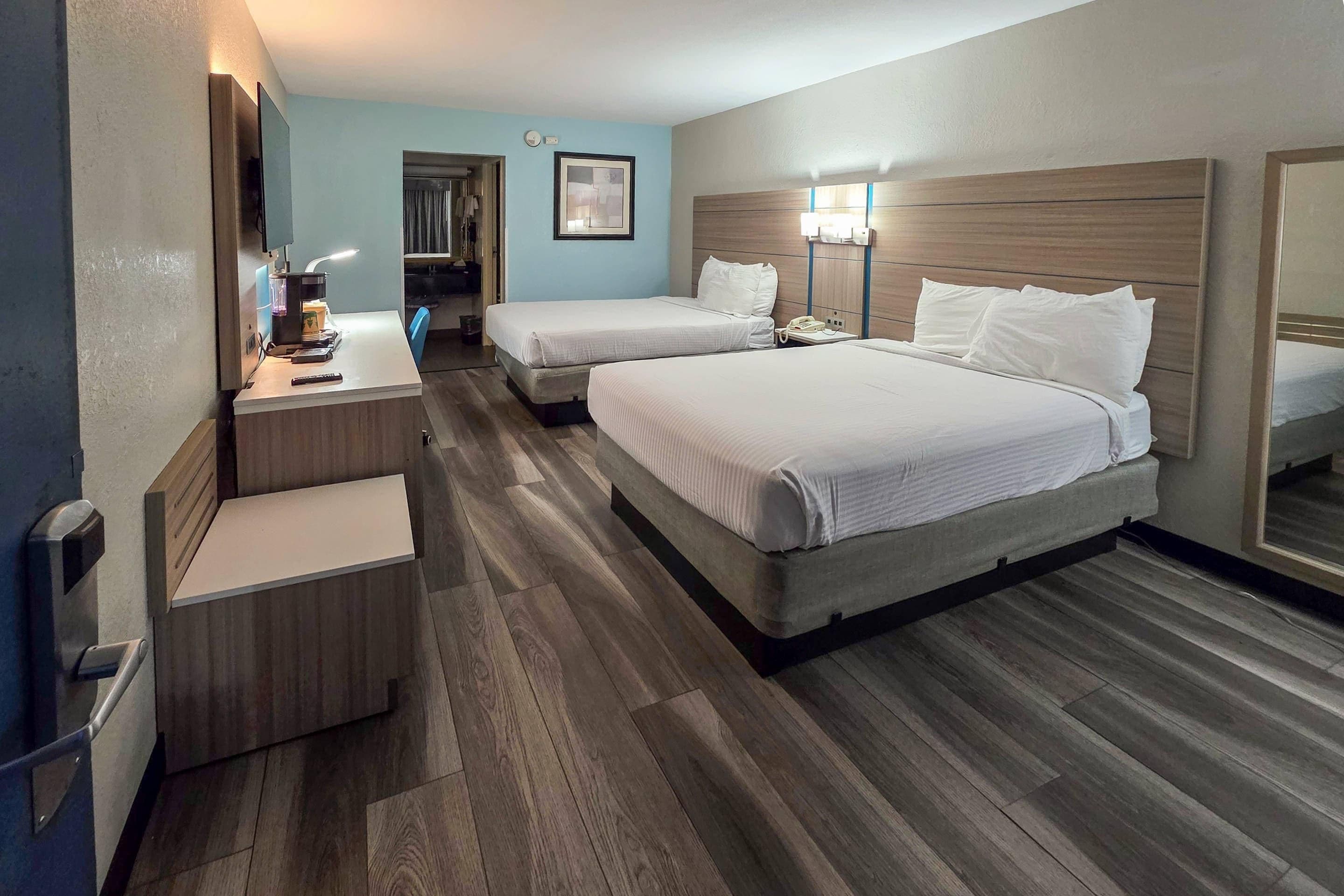 A hotel room with two beds, a desk with a lamp, a mirror, and a wooden floor.