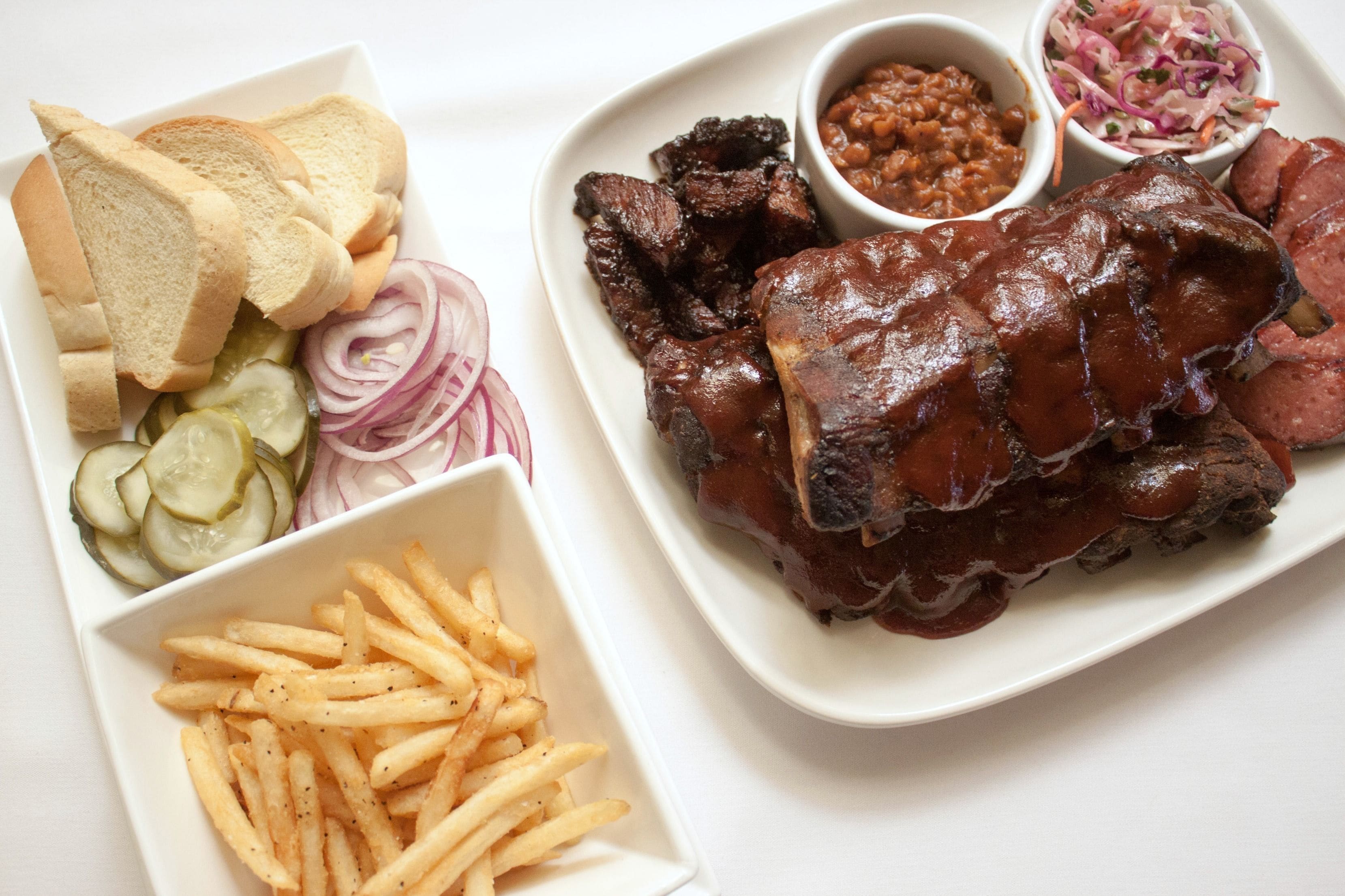 A plate of barbecued ribs, French fries, pickles, onions, and bread.