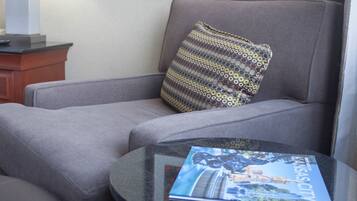 Una habitación de hotel moderna con un sofá gris, una mesa redonda con una revista, una lámpara y un cuadro abstracto enmarcado en la pared.