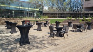 Un patio al aire libre con mesas y sillas redondas, un muro de piedra y un edificio moderno con ventanas de vidrio al fondo.