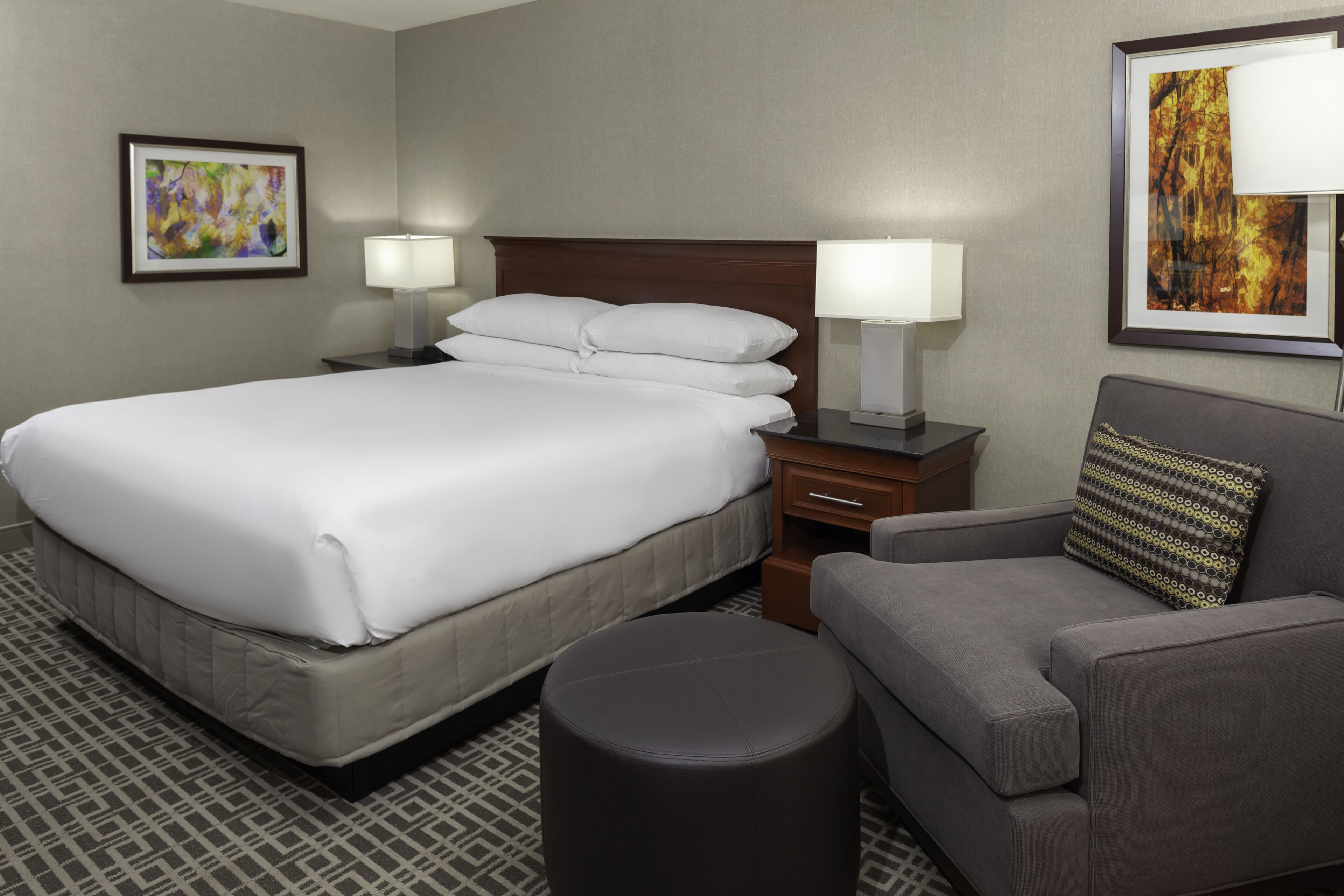 Una habitación de hotel con una cama grande, dos mesitas de noche con lámparas, un sillón gris y un suelo con estampado.