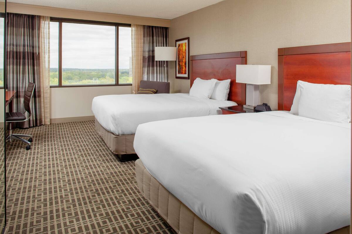 A hotel room with two beds, a desk, a chair, and a large window with a view.