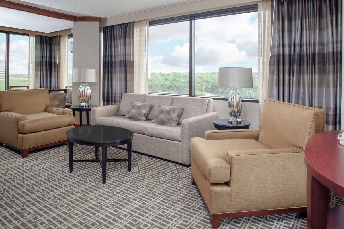 A hotel room with a sofa, armchairs, a coffee table, and a view of the outdoors.