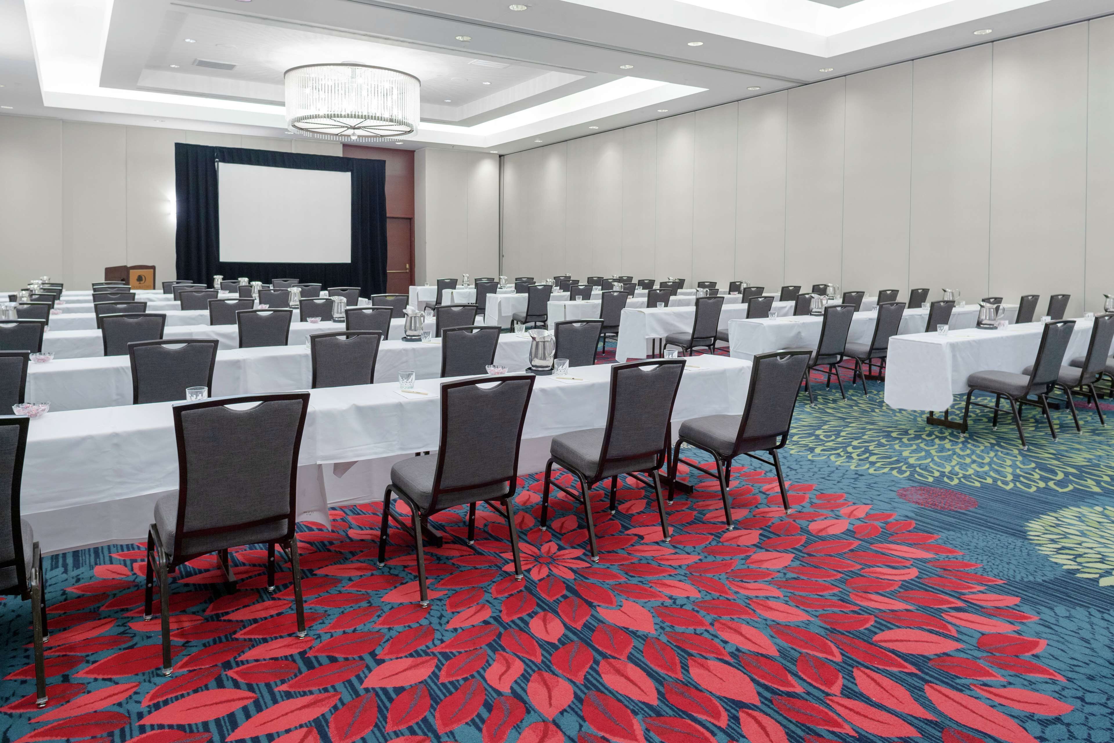 A conference room with rows of tables and chairs, a projector screen, and a carpeted floor.