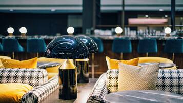 A modern bar area with a row of blue bar stools and a seating area with patterned cushions and a central table with a lamp.