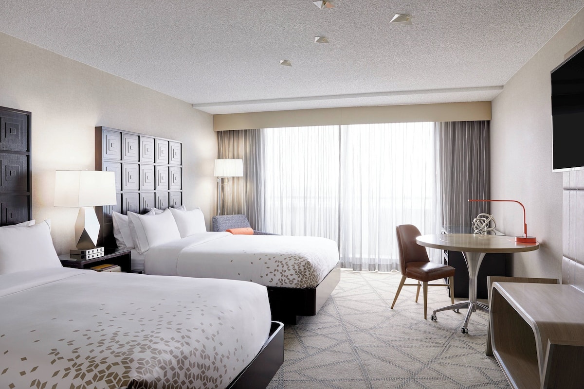 A hotel room with two beds, a desk, a chair, a TV, and a large window with curtains.