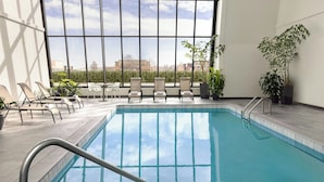 A swimming pool with lounge chairs and potted plants, large windows with a view of buildings, and a tiled floor.