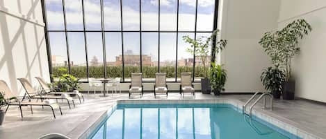 A swimming pool with lounge chairs and potted plants, large windows with a view of buildings, and a tiled floor.