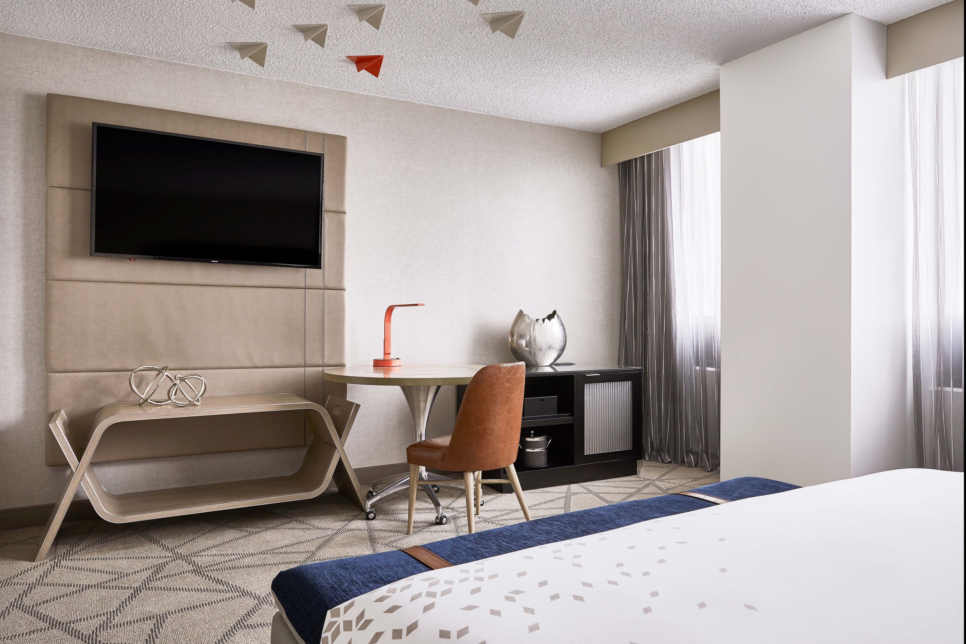 A hotel room with a bed, a desk with a chair, a television mounted on the wall, and a window with curtains.
