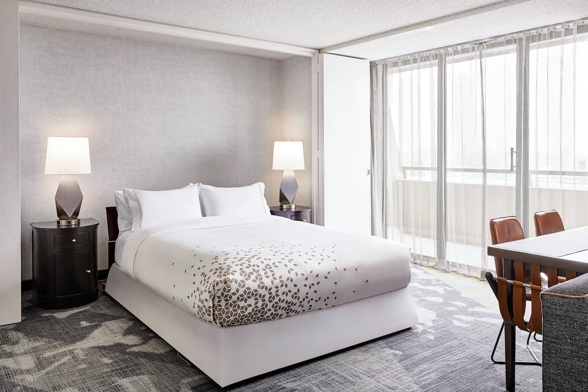 A hotel room with a large bed, two bedside lamps, a dining table with chairs, and a view through sheer curtains.