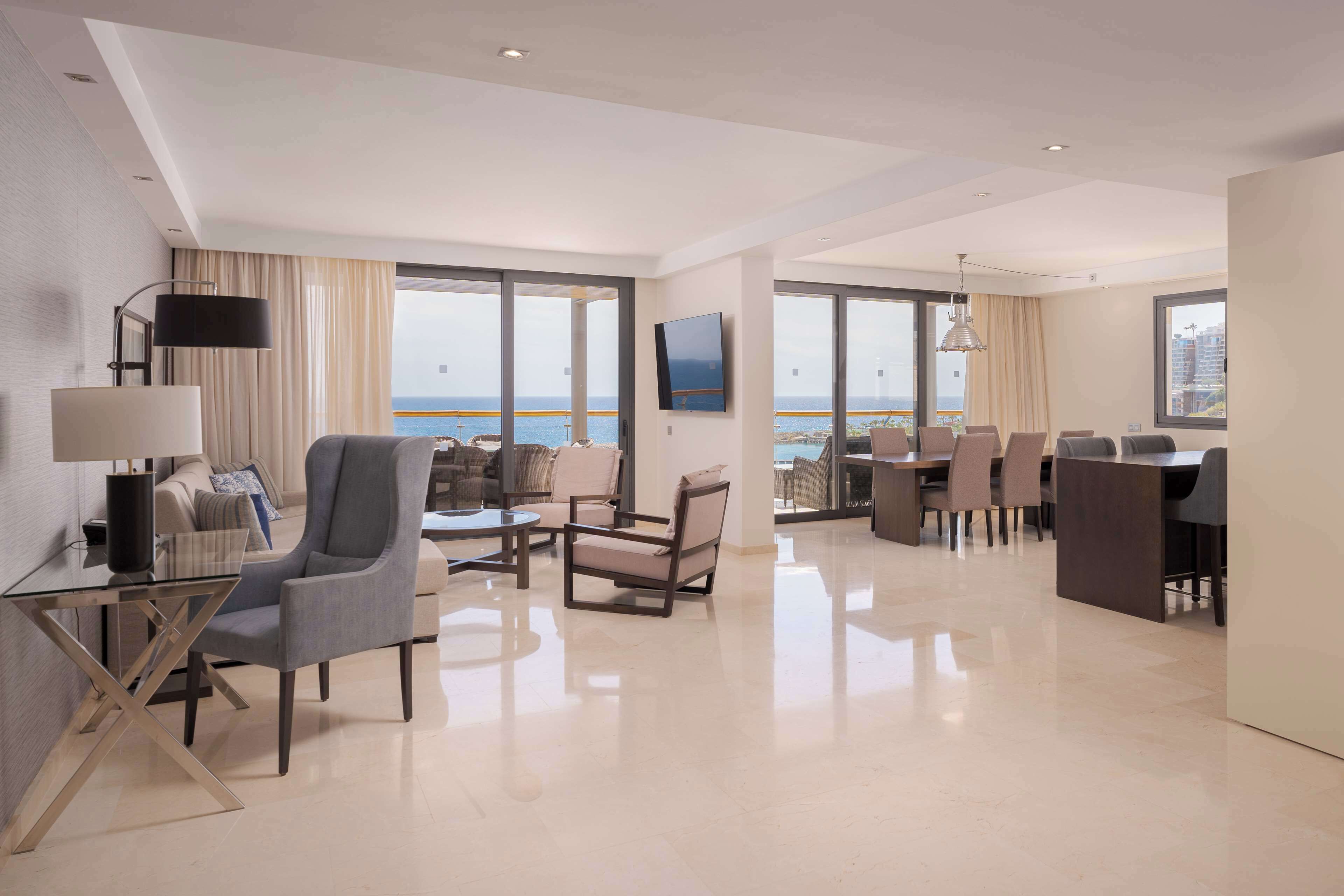 Ein modernes Hotelzimmer mit Essbereich, großen Fenstern mit Meerblick und einem an der Wand befestigten Flachbildfernseher.