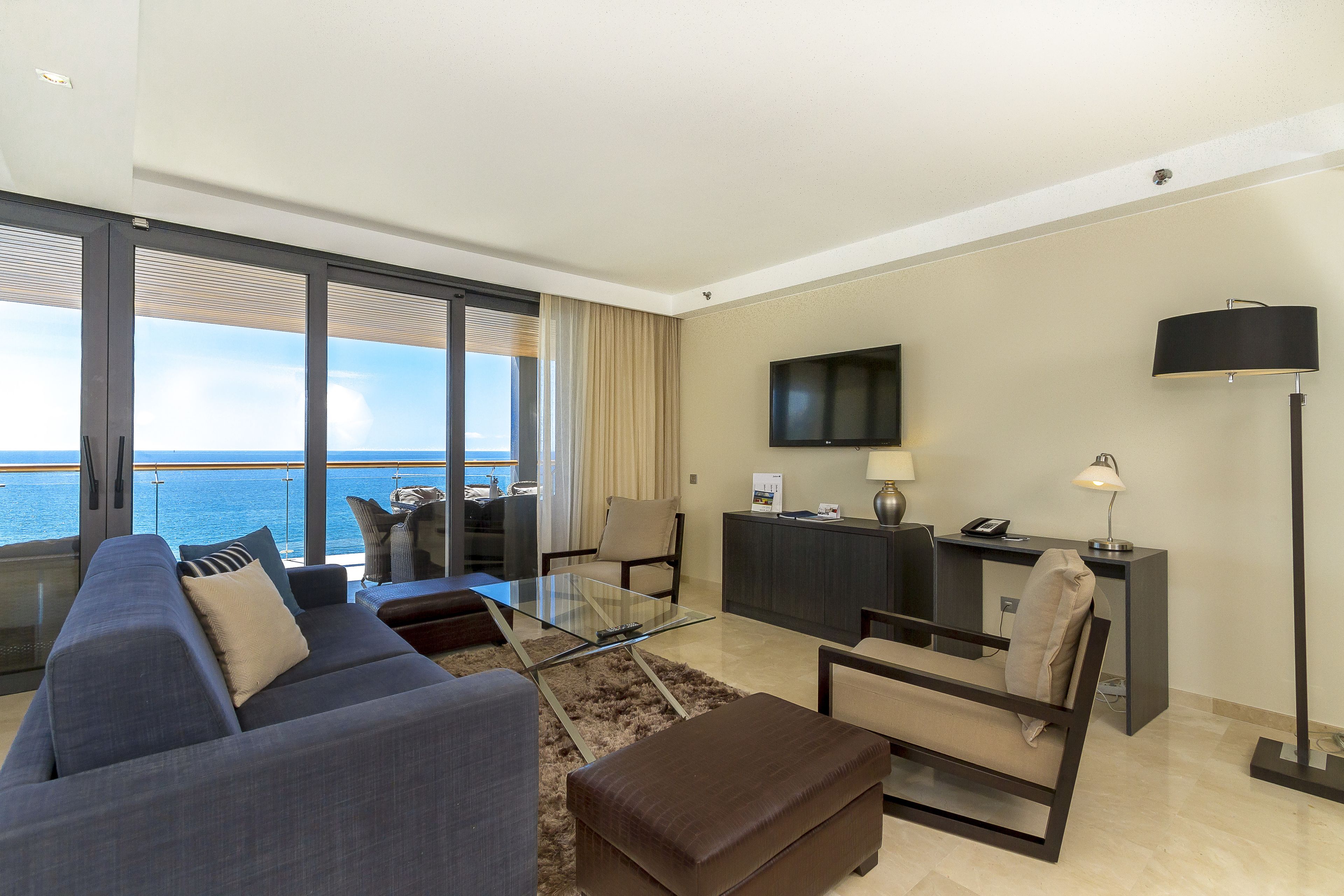 A modern living room with a sofa, armchair, ottoman, and a glass coffee table. There is a flat-screen TV mounted on the wall, a desk with a lamp, and a view of the ocean through large windows.