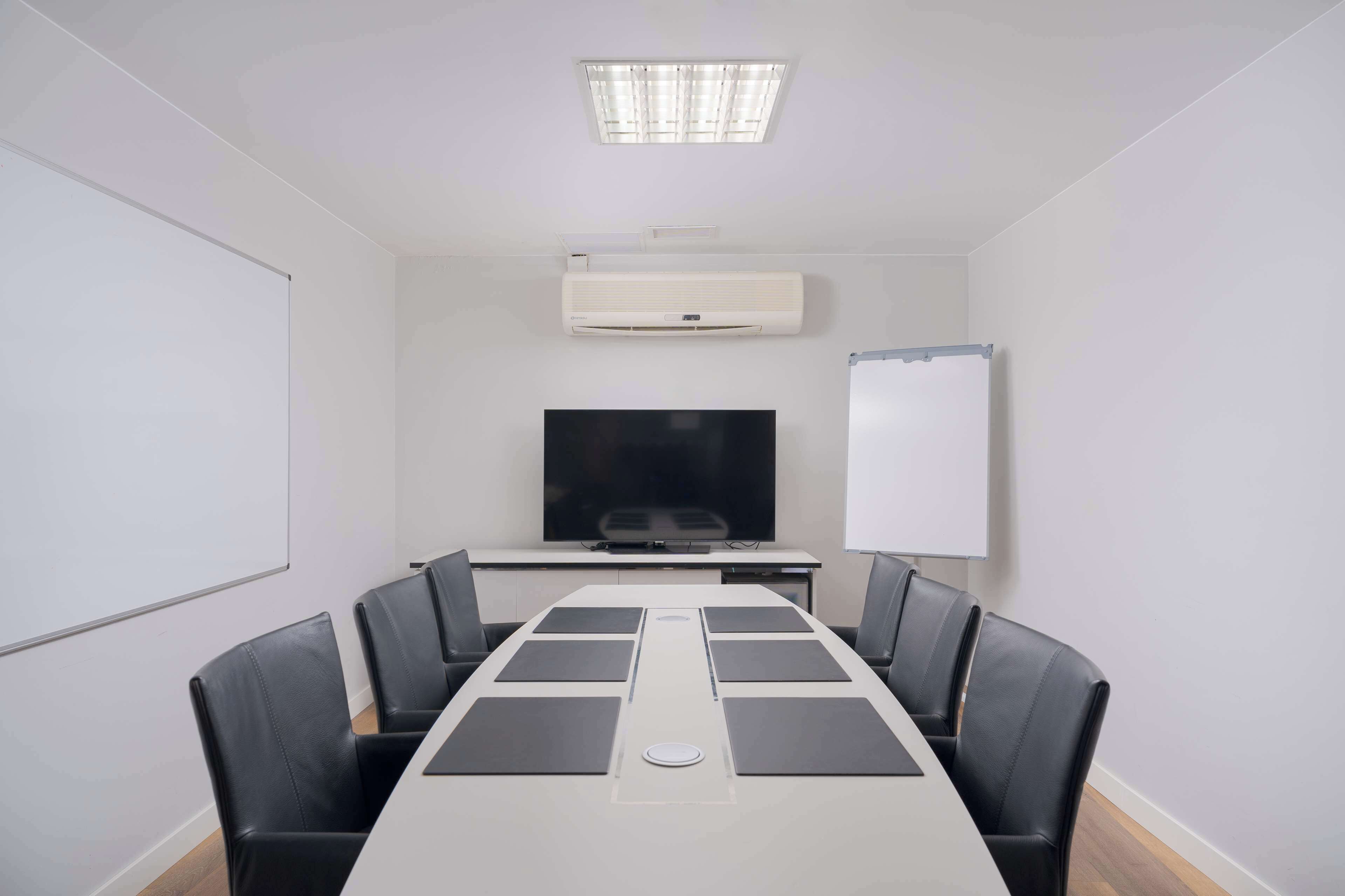 Una sala de conferencias con una larga mesa, sillas negras, un televisor de pantalla plana, una pizarra blanca y un equipo de aire acondicionado.