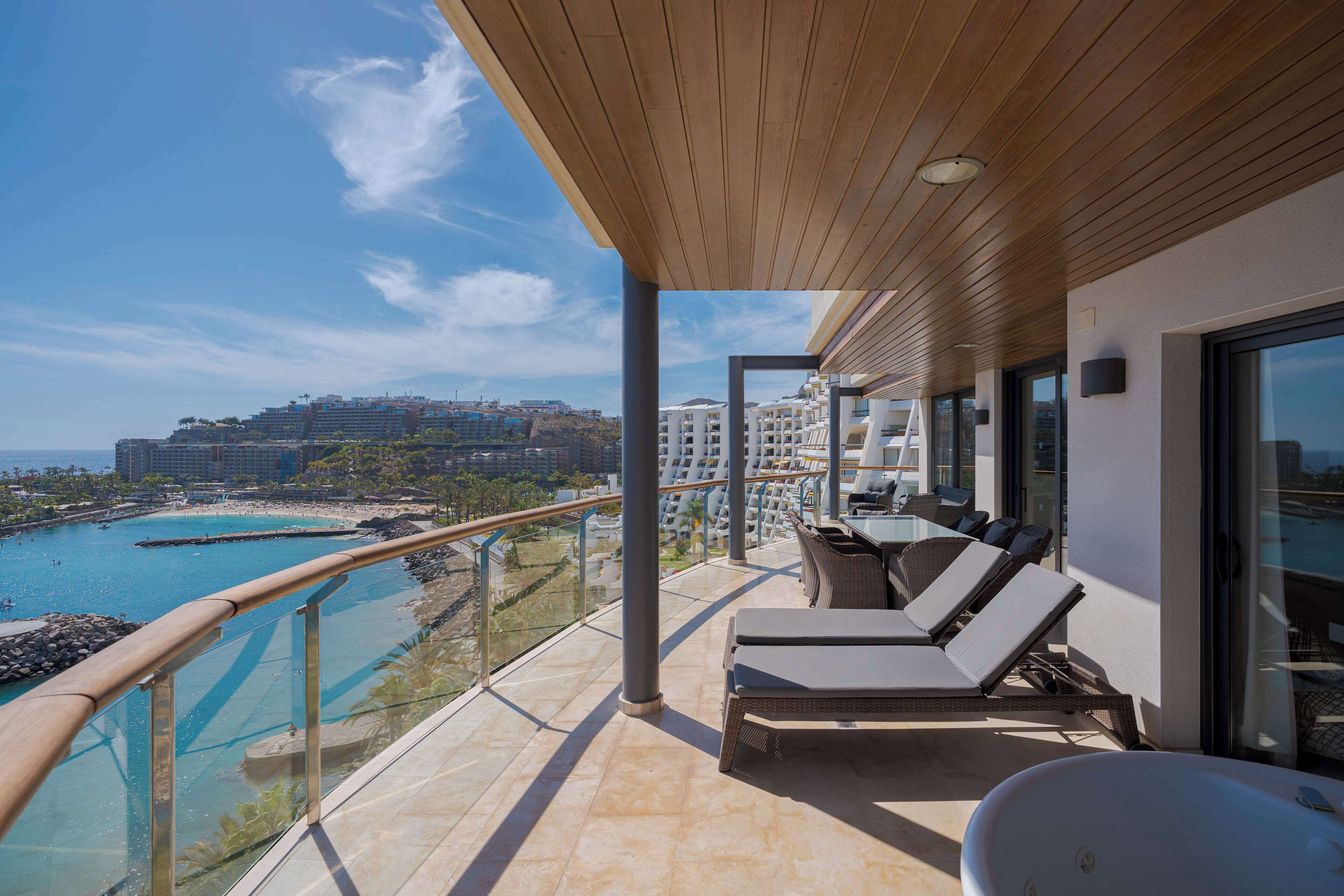 A balcony with a view of a beach and ocean, furnished with lounge chairs and a sun lounger.