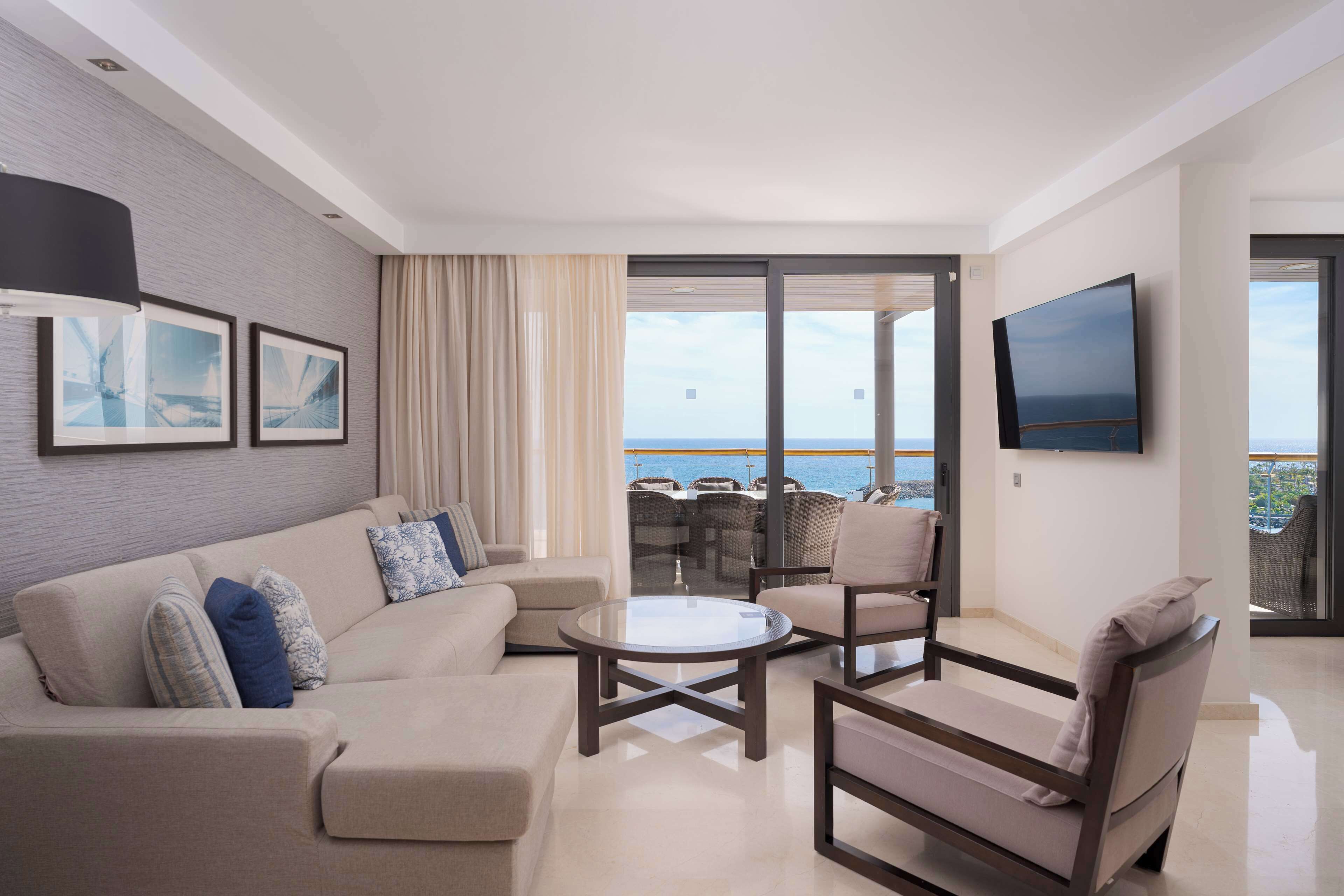 Een moderne woonkamer met een bank, fauteuils, een salontafel en een televisie. Er is een groot raam met uitzicht op zee.