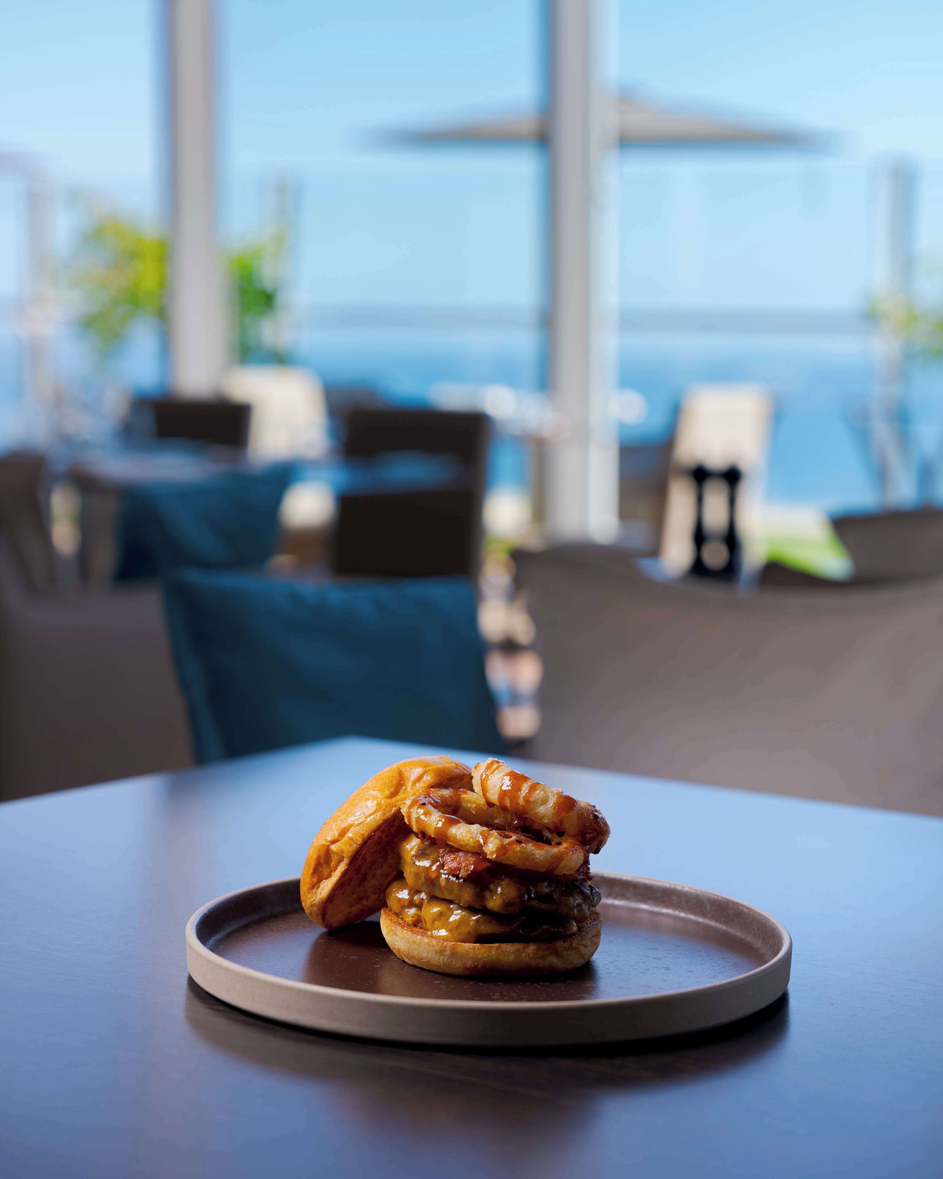 A burger with onion rings on a plate on a table in a restaurant with a view of the ocean.