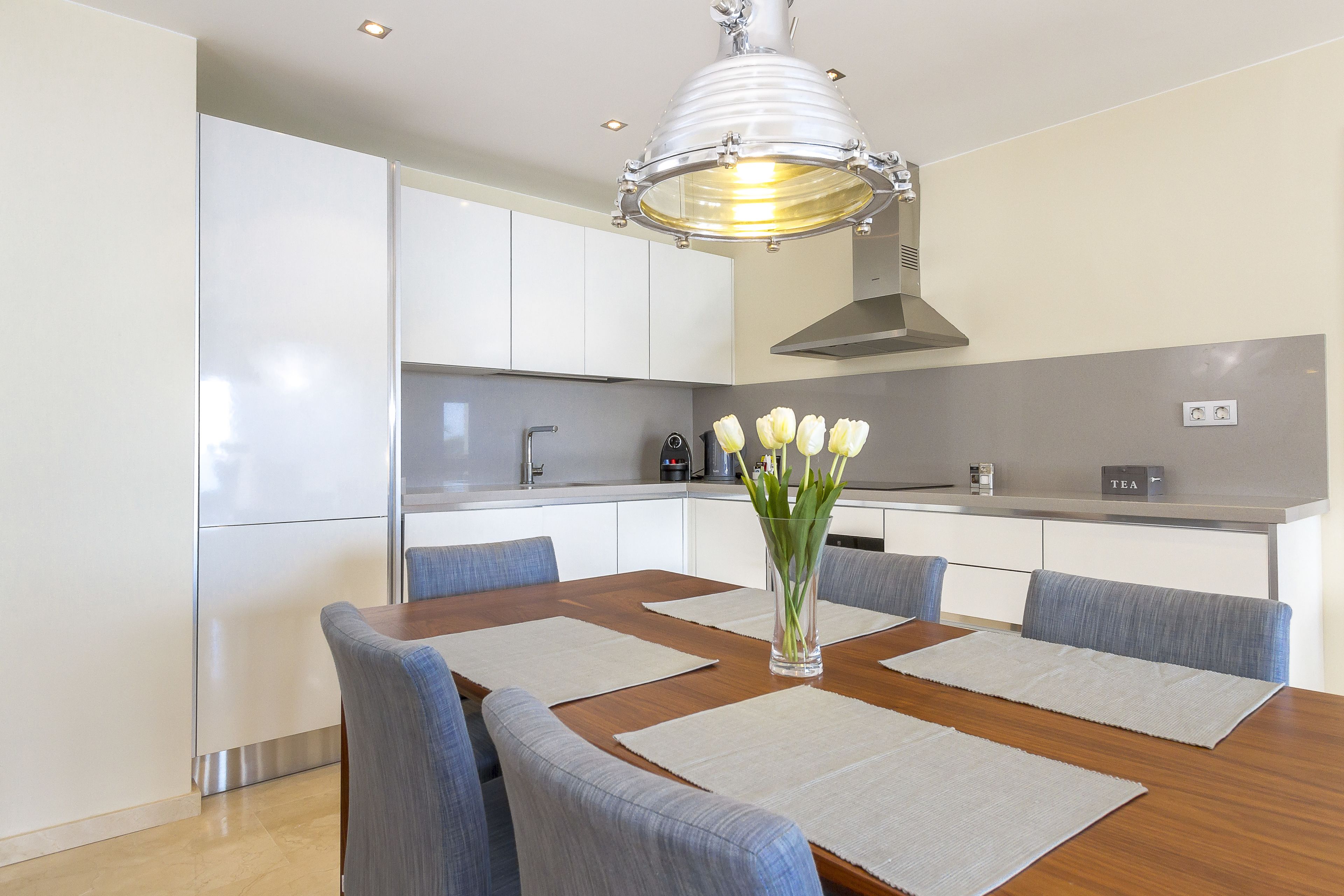 Una cocina moderna con armarios blancos, una mesa de comedor de madera con sillas azules y un jarrón con flores blancas.