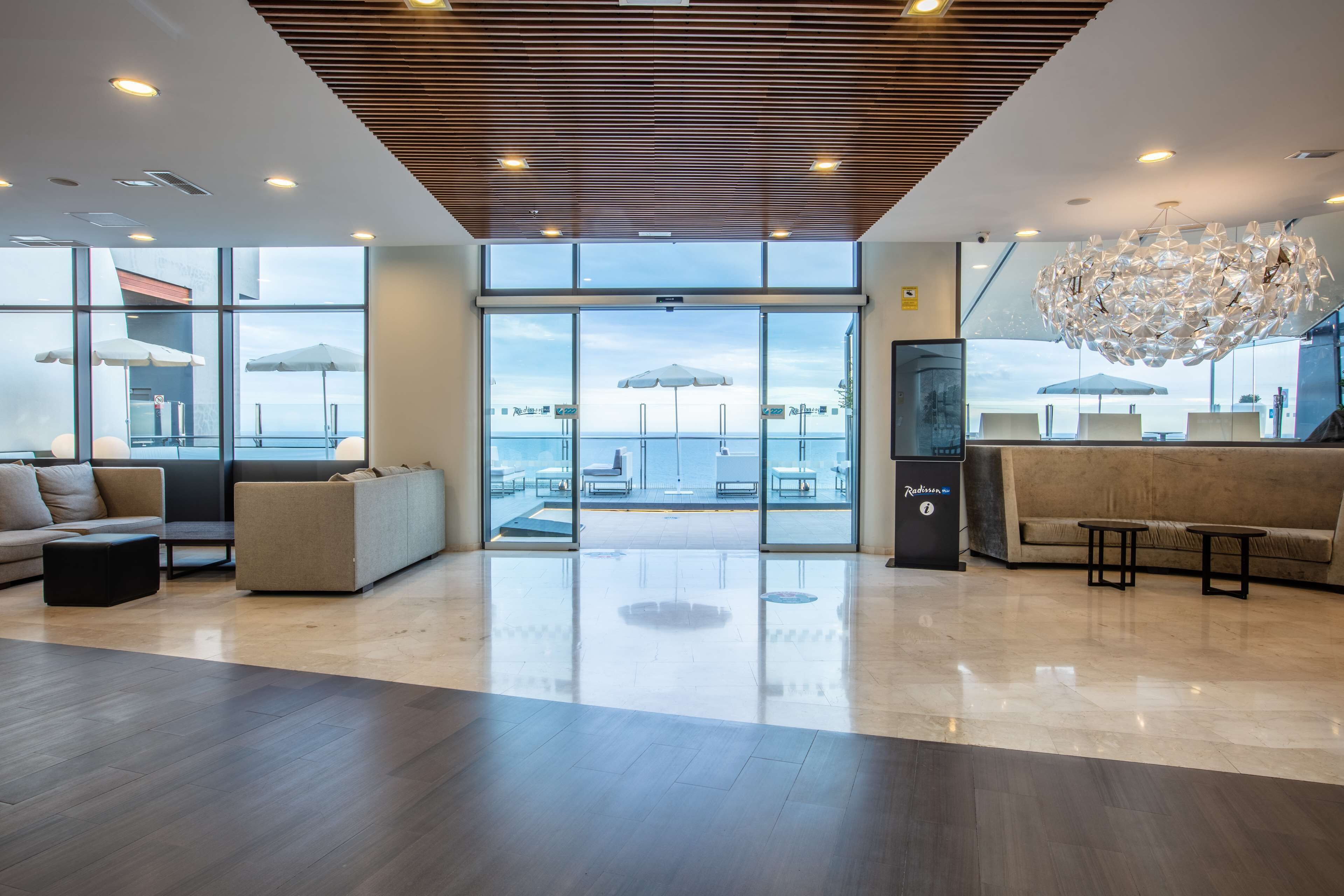 A spacious lobby with a modern design, featuring a wooden ceiling, large glass doors leading to a terrace with outdoor seating, and a central chandelier.
