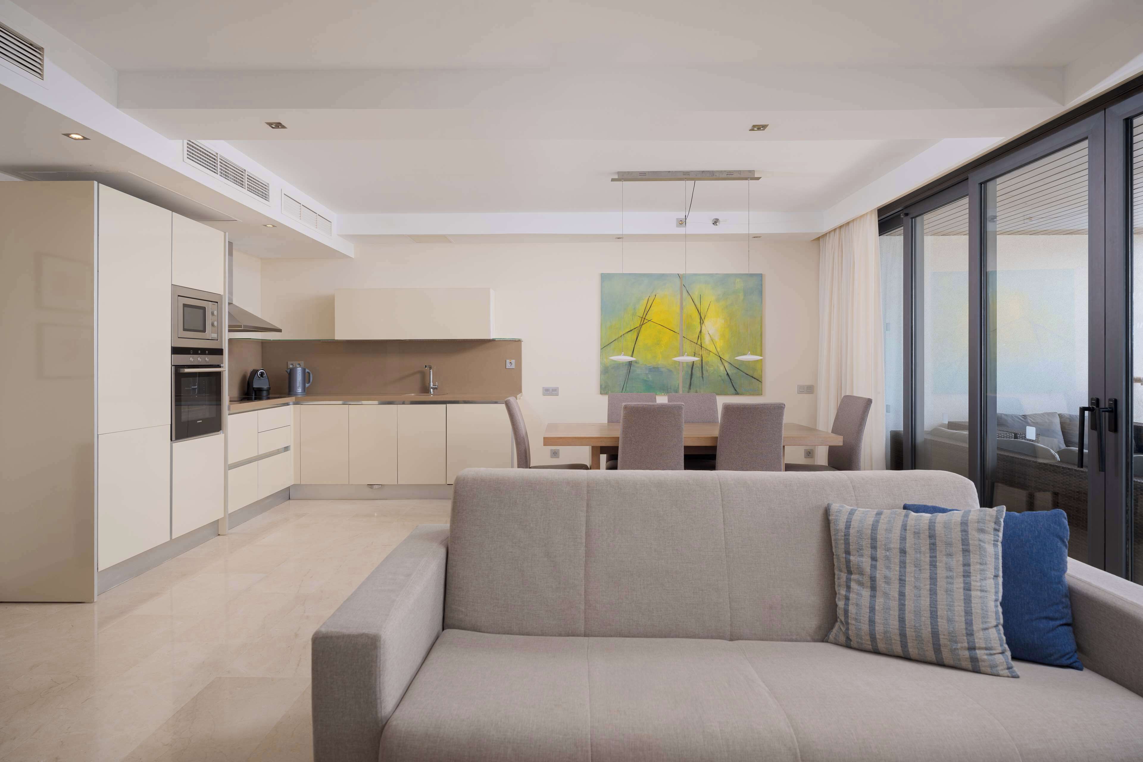 A modern living room with a sofa, a dining table with chairs, a kitchen area with cabinets and appliances, and a large window with a view.