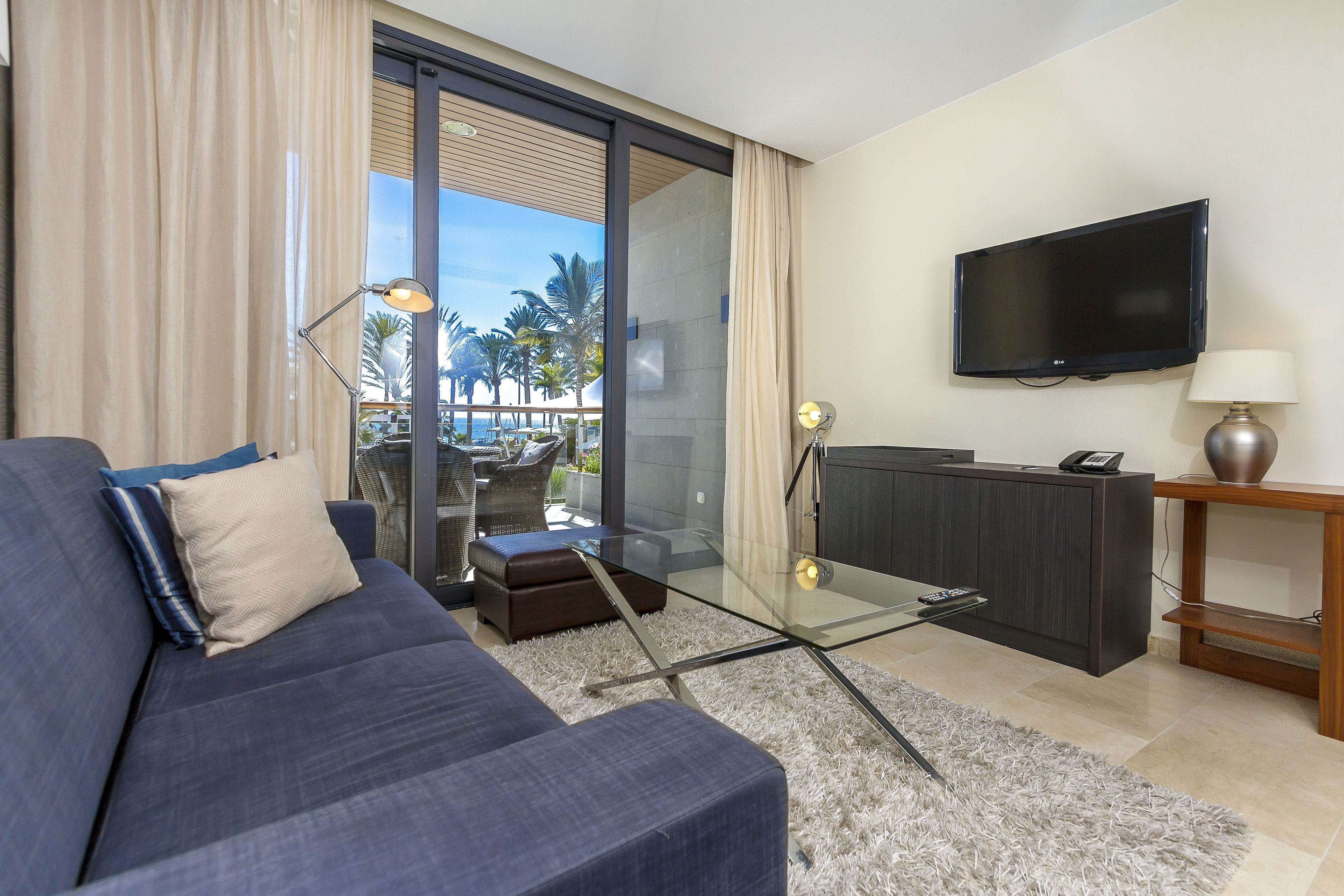 A modern living room with a sofa, a glass coffee table, a TV, and a view of palm trees through a sliding glass door.