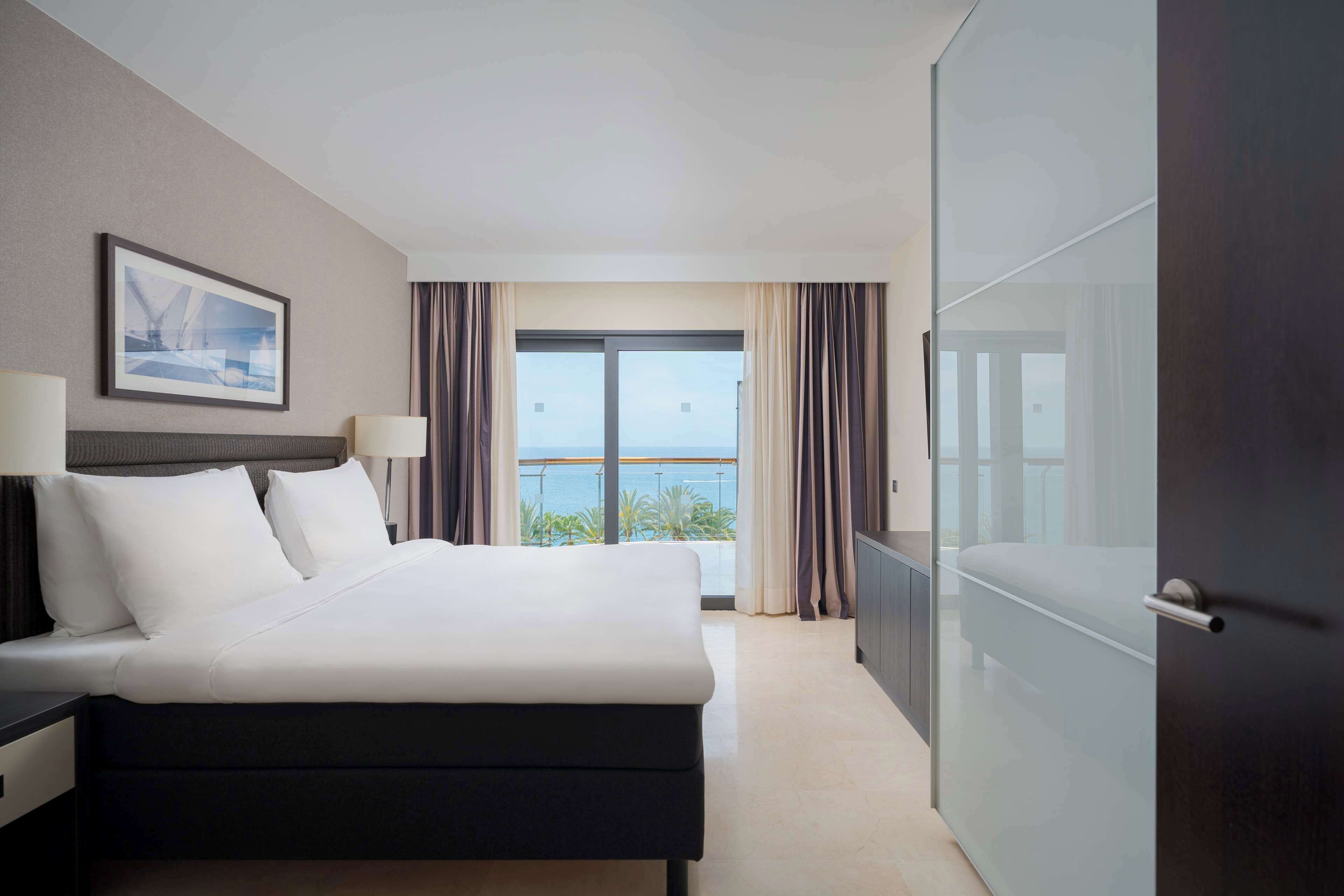 Habitación de hotel con una cama grande, una mesita de noche, una lámpara, un cuadro en la pared, una puerta corrediza de vidrio que da a un balcón con cortinas y un armario de color oscuro.