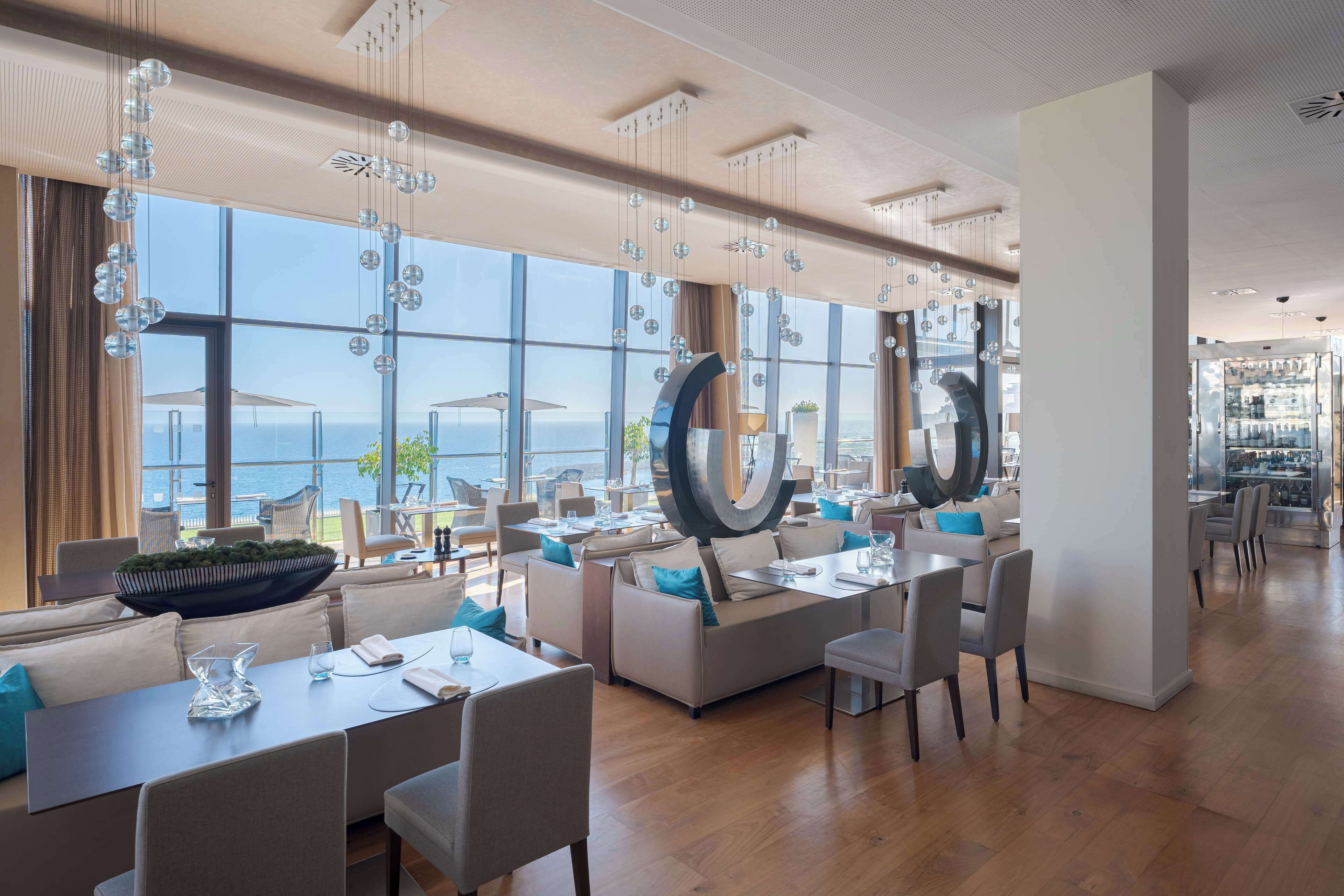 A modern dining area with large windows, a view of the sea, and contemporary furniture.