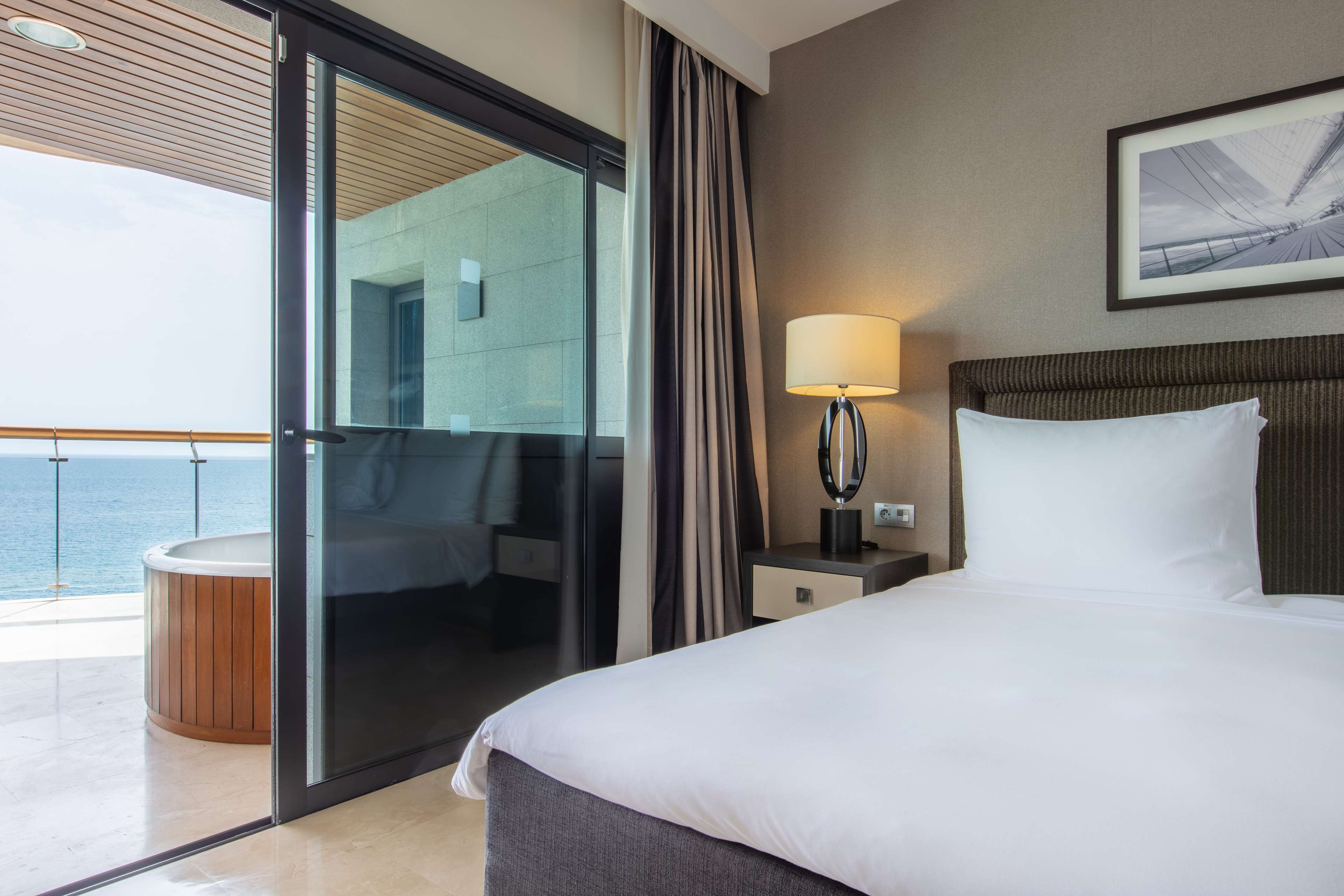 A hotel room with a bed, a nightstand, a lamp, a large window with curtains, and a view of the sea.