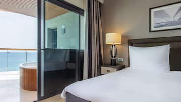 A hotel room with a bed, a nightstand, a lamp, a large window with curtains, and a view of the sea.