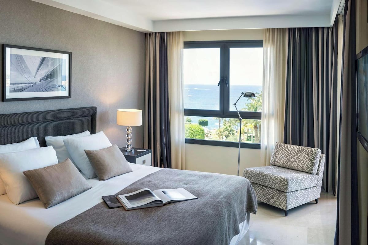 Ett hotellrum med en säng, en stol, en tv, ett fönster med gardiner och en lampa.