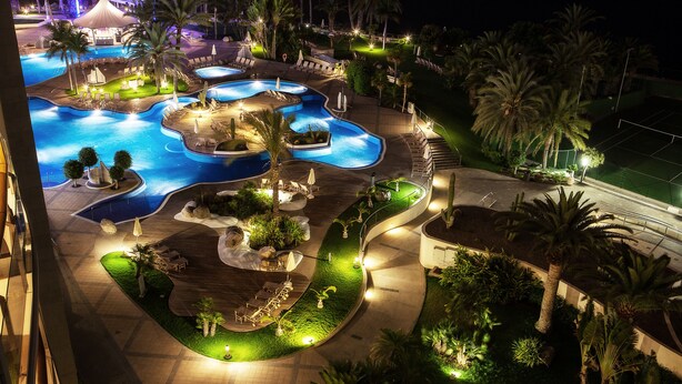 Vista nocturna de un resort con varias piscinas, palmeras y zonas de estar al aire libre.