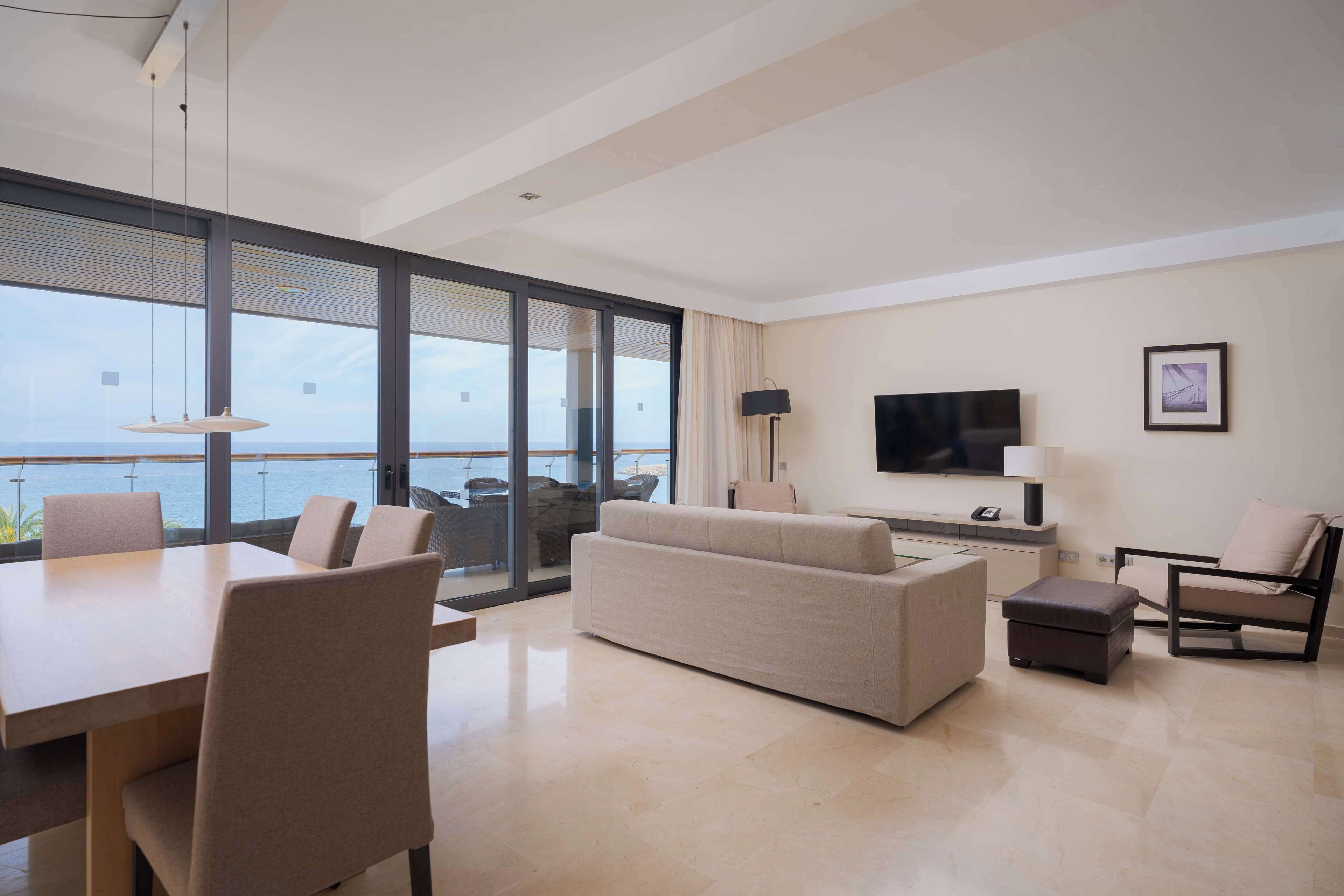 Un salón moderno con un ventanal grande, un televisor de pantalla plana, un sofá, una mesa de comedor y un balcón con vista al mar.