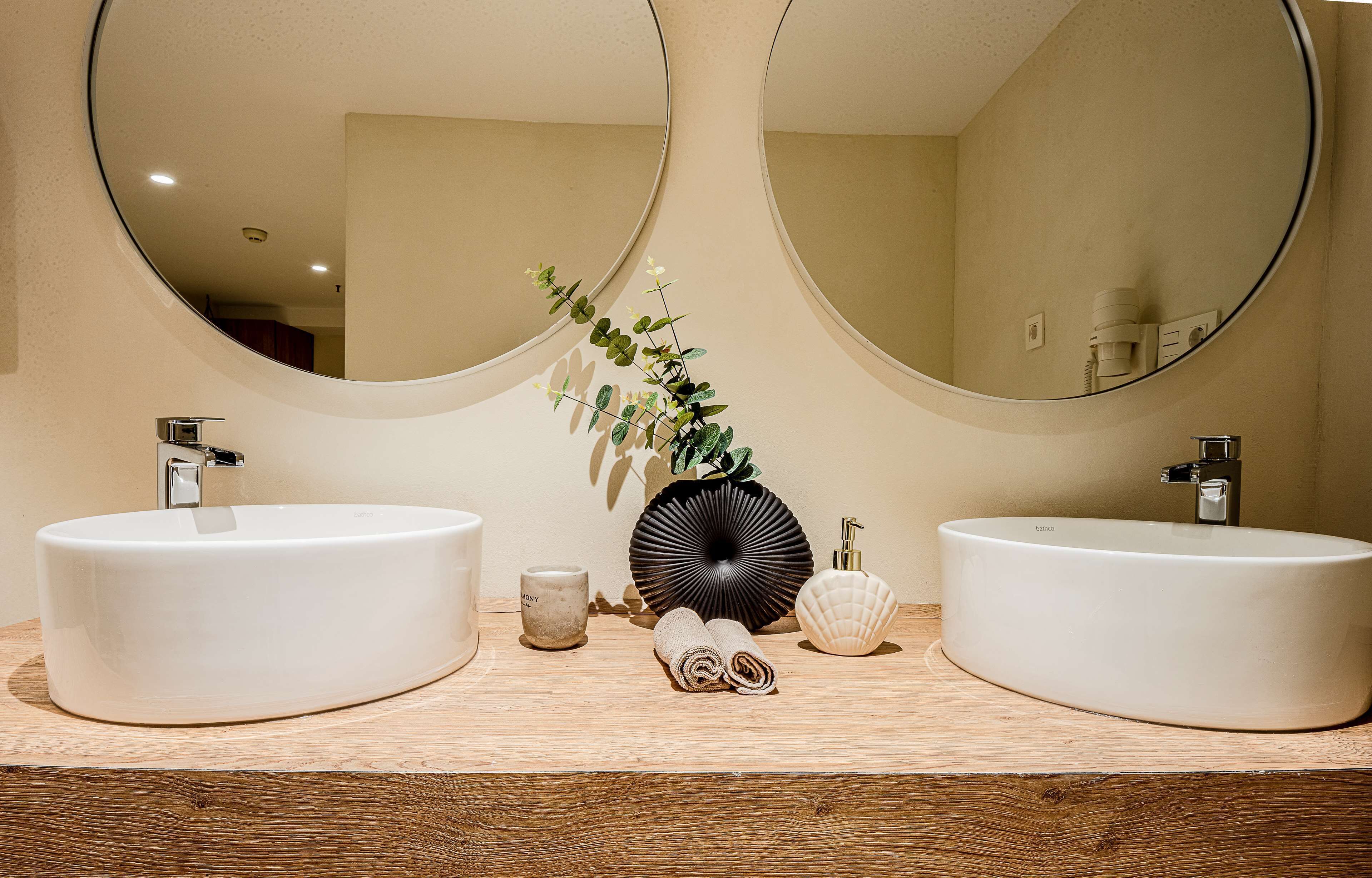 Un baño con dos lavamanos blancos y redondos, un mueble de tocador de madera y dos espejos grandes.
