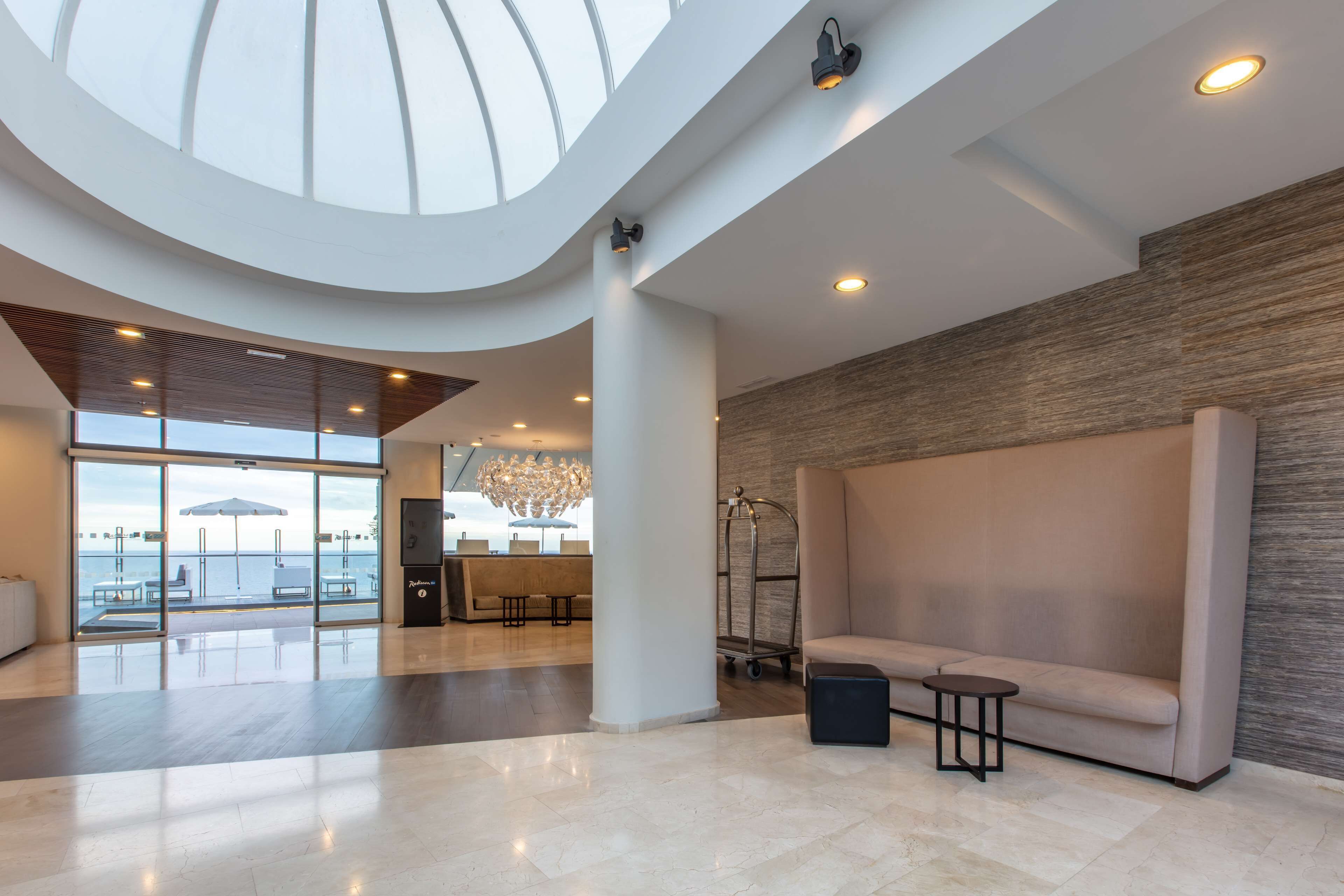 A spacious lobby with a high ceiling, large windows offering a view of the sea, a seating area with a sofa and a small table, and a reception desk in the background.