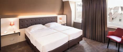 Una cama doble con cabecera capitonada, mesitas de noche con lámparas, una silla roja y vistas de edificios a través de una ventana con cortinas.