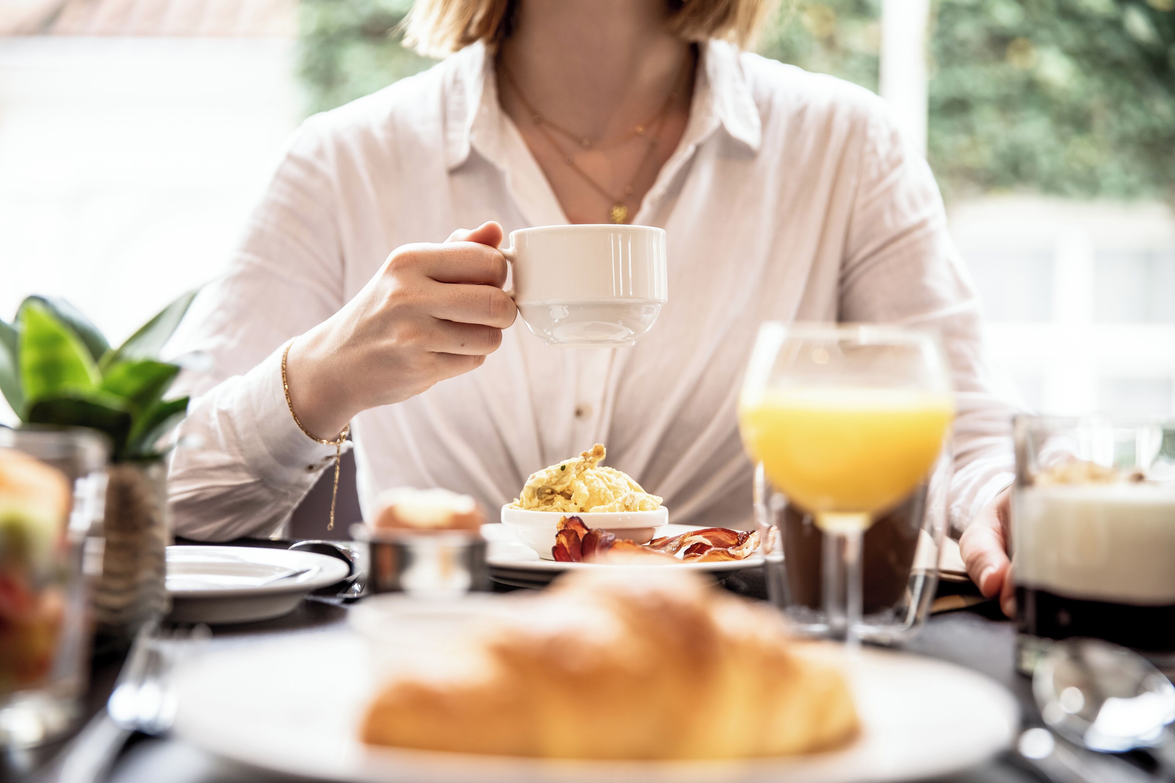 A person holding a white cup at a dining table with breakfast items including eggs, bacon, and a croissant.