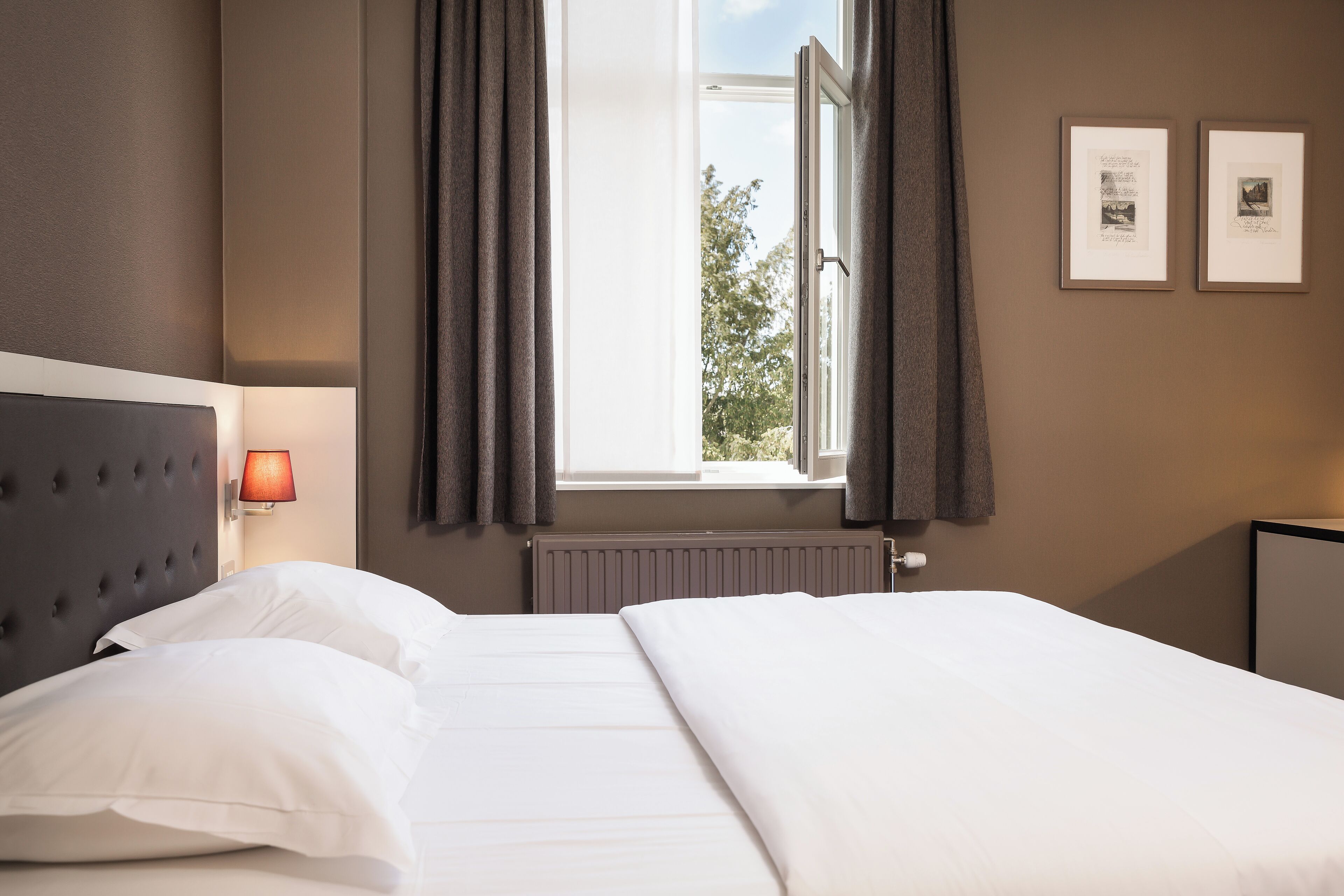 A hotel room with a bed, a window with curtains, a wall-mounted lamp, and two framed pictures.