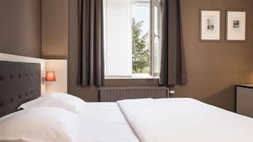 A hotel room with a bed, a window with curtains, a wall-mounted lamp, and two framed pictures.