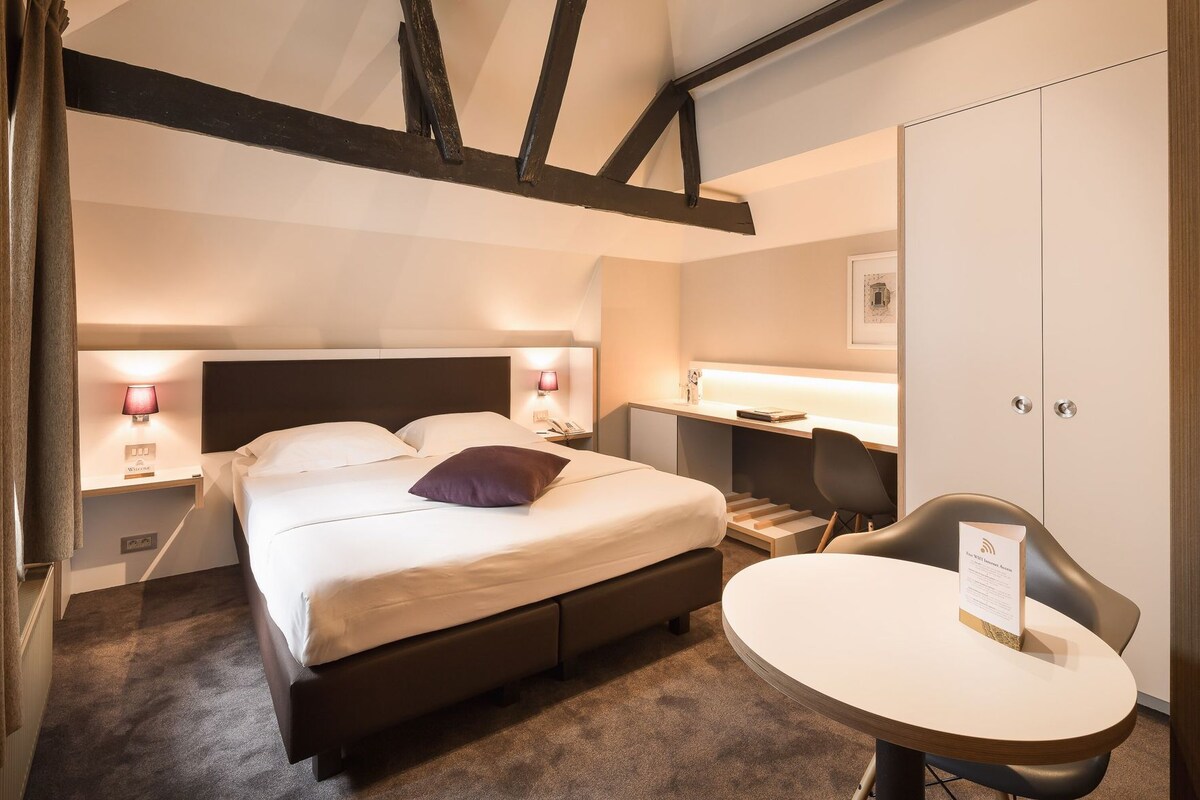 Una habitación de hotel con una cama, lámparas de noche, un escritorio, una silla y una mesita.