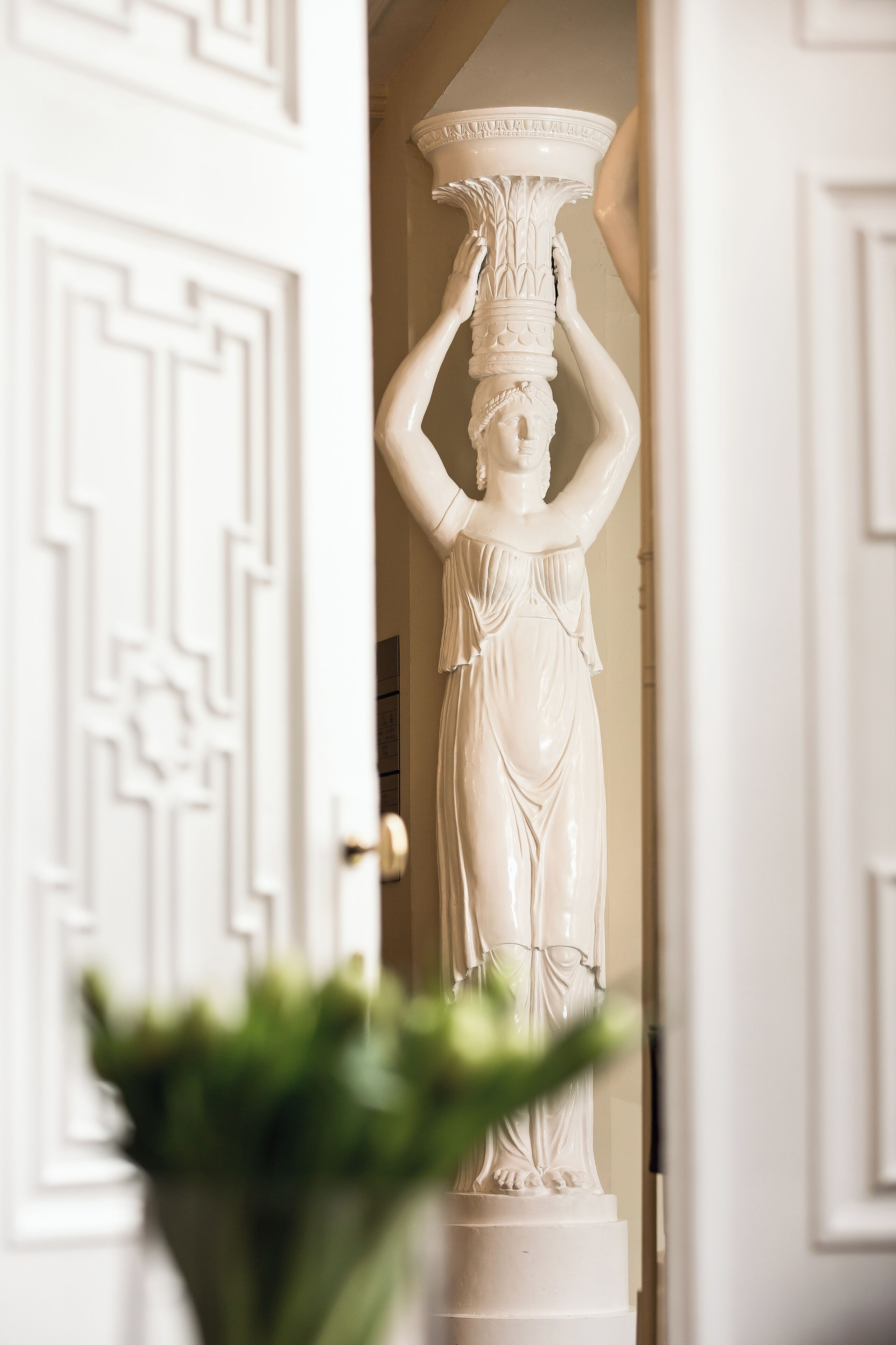 Eine weiße Statue einer Frau, die eine Schale auf dem Kopf balanciert, steht in einer Türöffnung mit dekorativem Türrahmen.