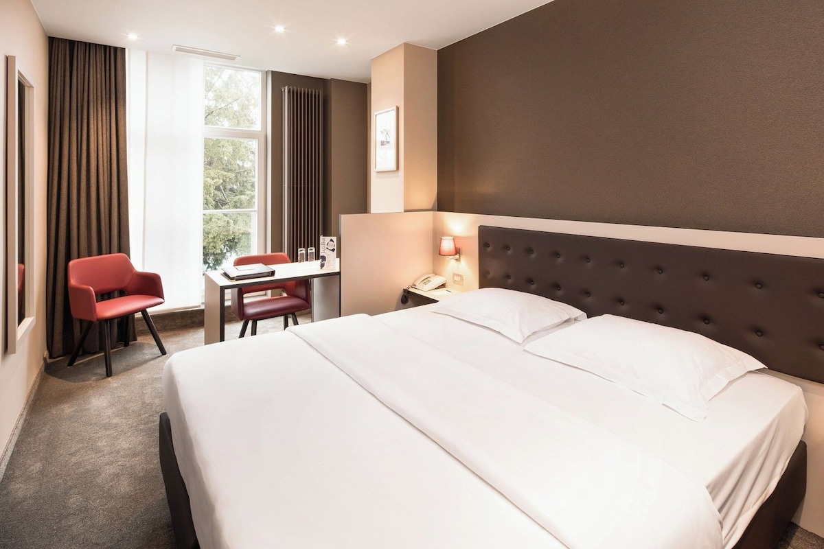 Habitación de hotel con una cama grande, un escritorio con silla, una ventana con cortinas y un radiador.
